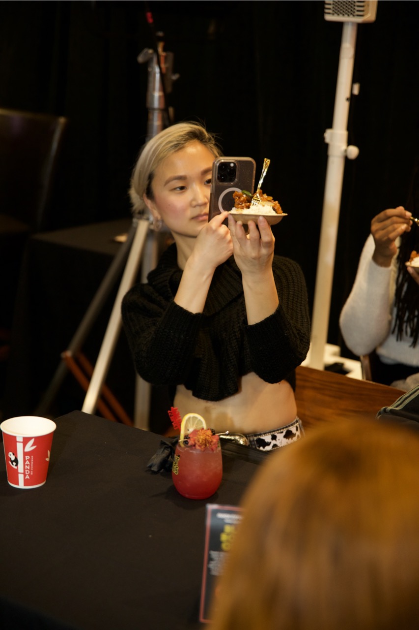 Young woman with short blonde hair taking a photo of a dessert with her phone, sitting at a table in a dark room.