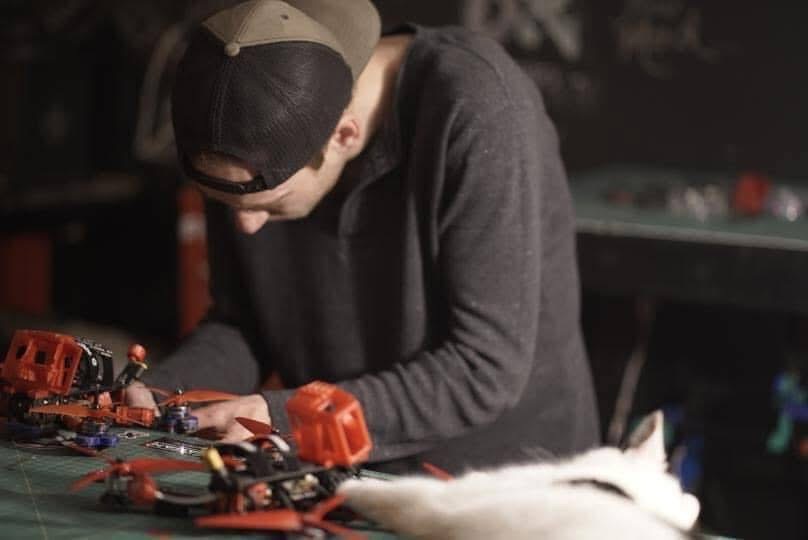 Person wearing a cap and glasses working on a table with small toy vehicles and objects, with a white animal nearby.