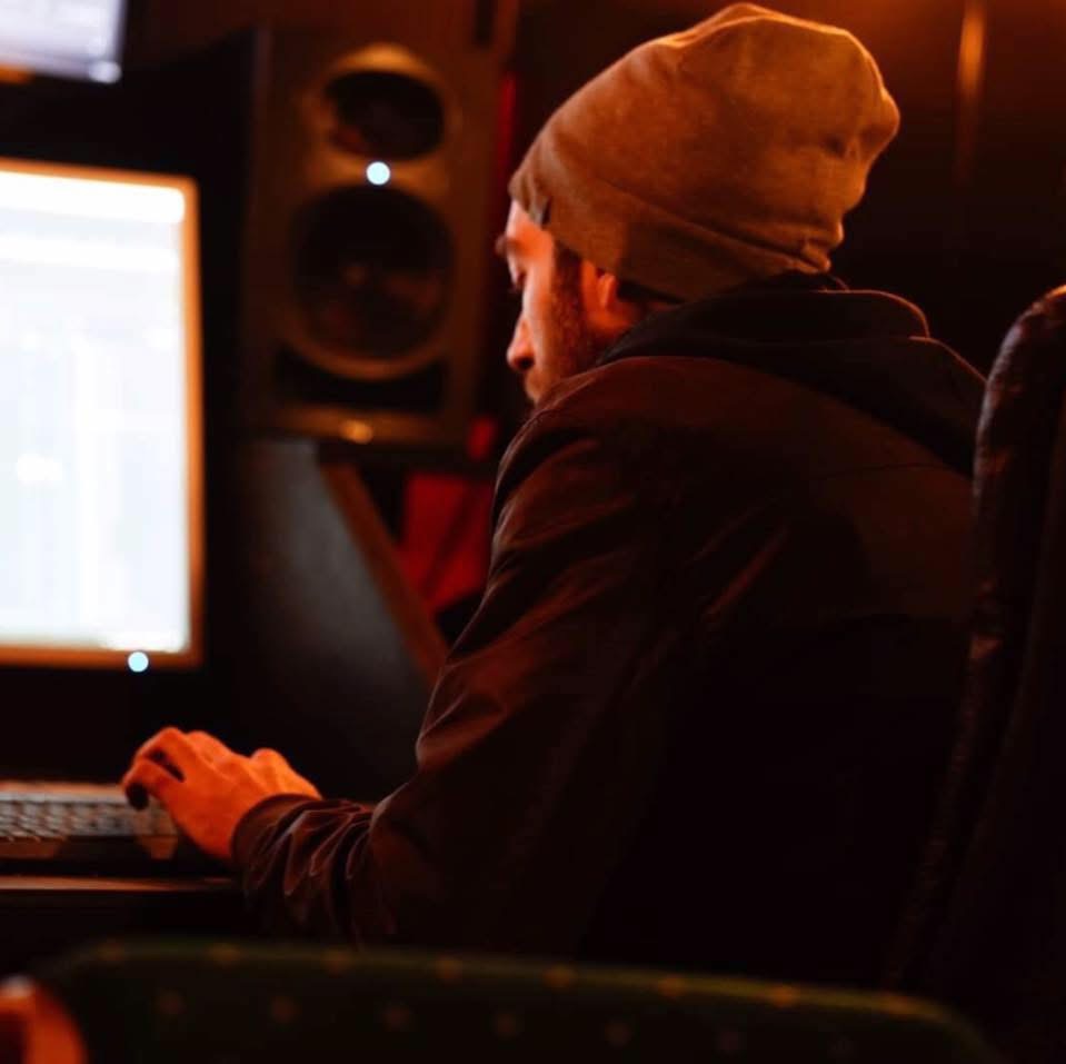 Person wearing a beanie and hoodie working on a computer in a dimly lit room with speakers.