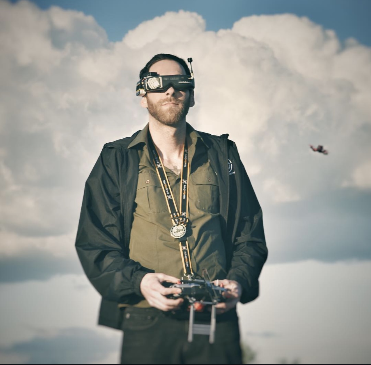 Man wearing goggles and a jacket, holding a remote control, standing outdoors with clouds in the sky.