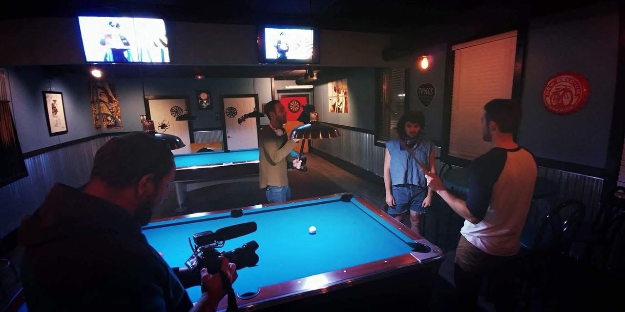 People playing pool in a dimly lit bar with multiple television screens on the wall.