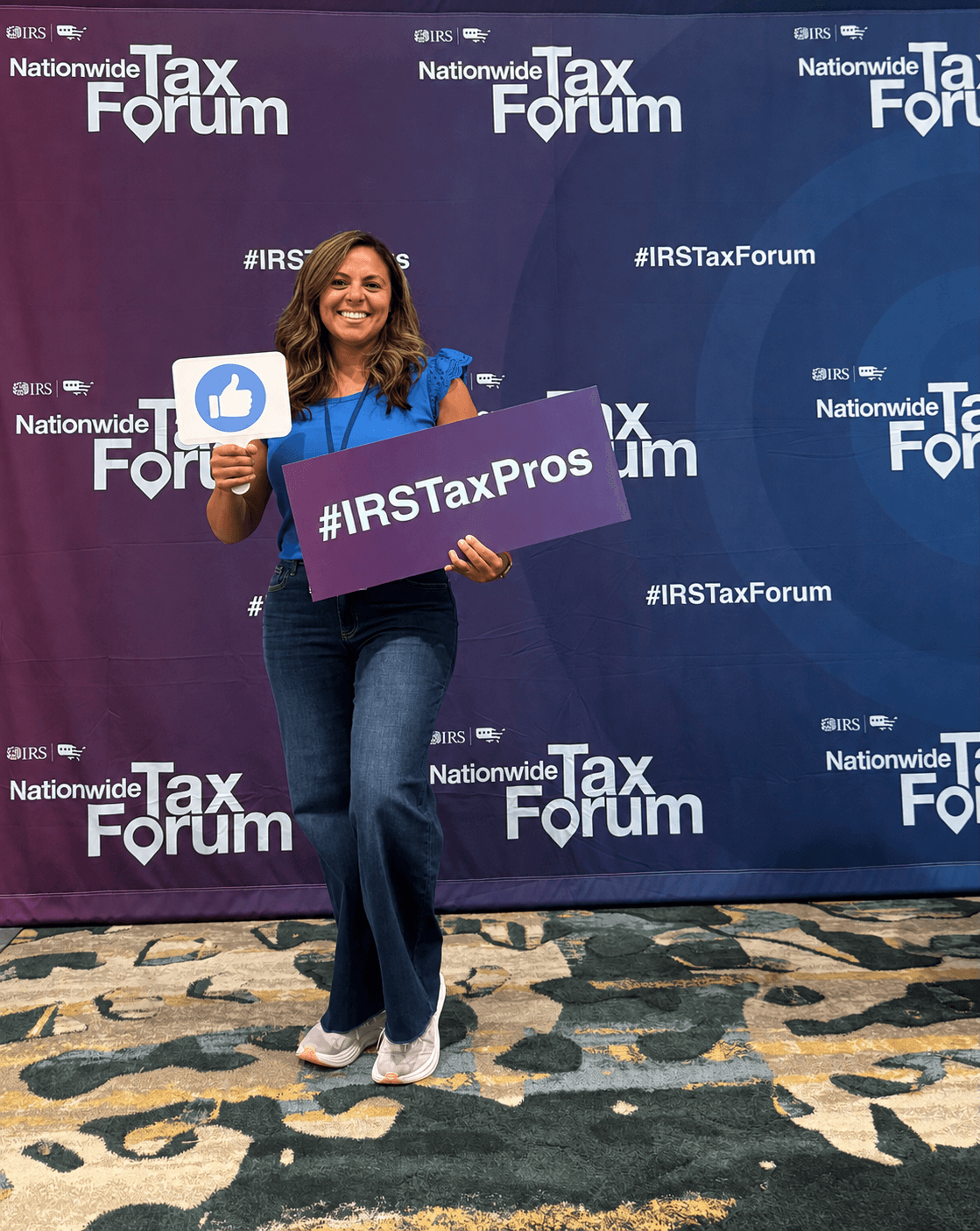 Woman holding a thumbs-up sign and a hashtag sign at a tax forum event, smiling, standing on a patterned floor.