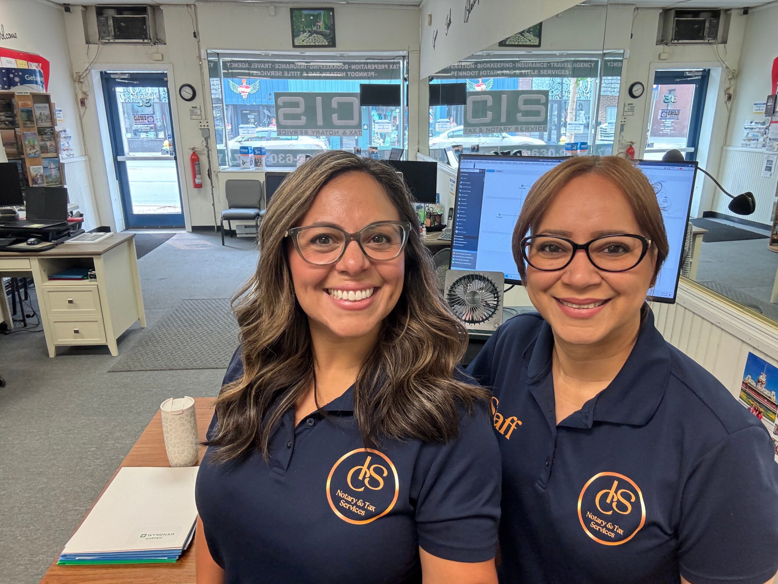 Two women wearing navy blue shirts with a logo, smiling in an office with computers and windows behind them.