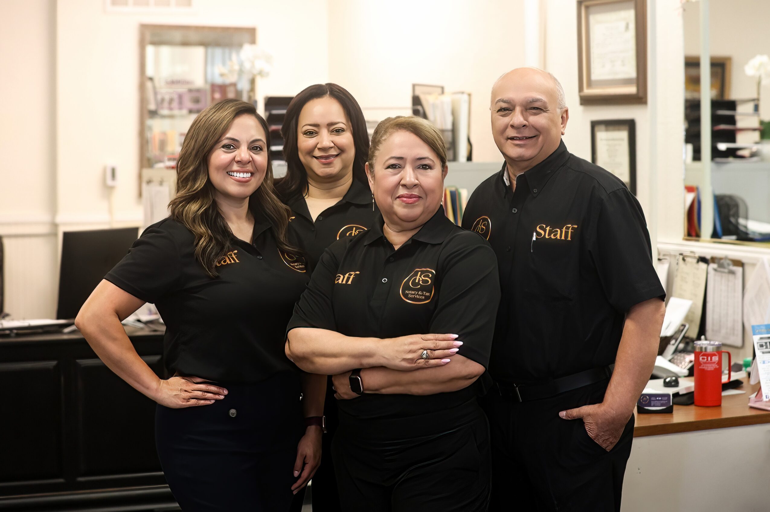 Four staff members in black uniforms standing in an office, smiling, with framed certificates on the wall behind them.