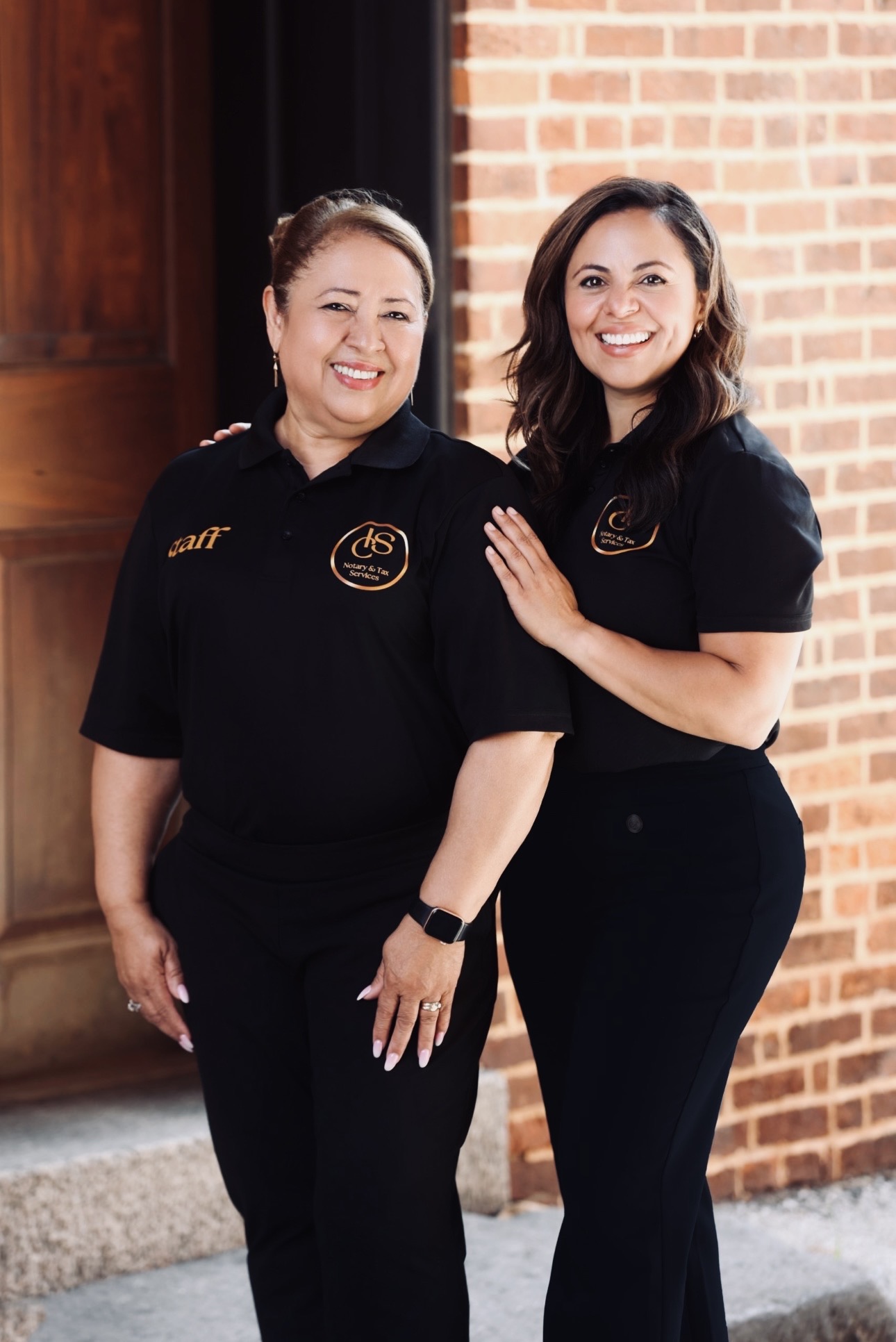 Two women in black uniforms standing outside near a brick wall, smiling, one with hand on the other's shoulder.