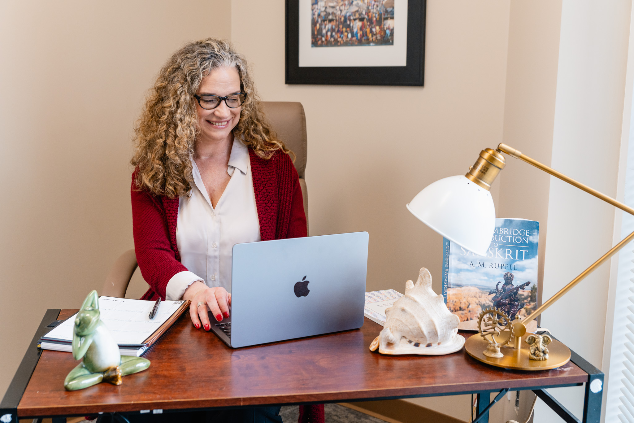 Woman with curly hair and glasses working on a laptop at a desk, smiling, in a well-lit room.
