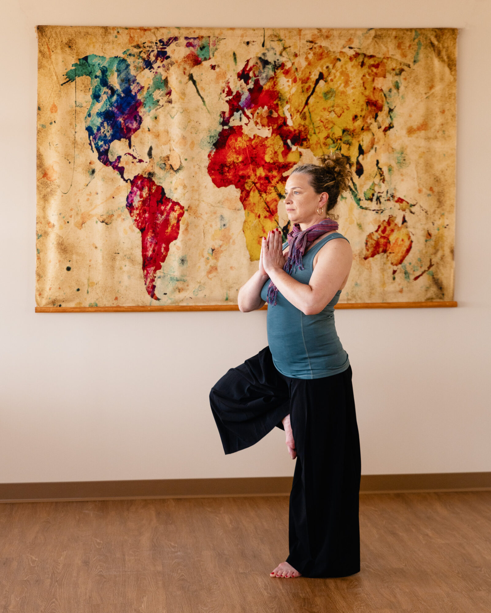 Woman standing with hands in prayer position in front of a colorful world map painting, holding a bag, barefoot, in a gallery.