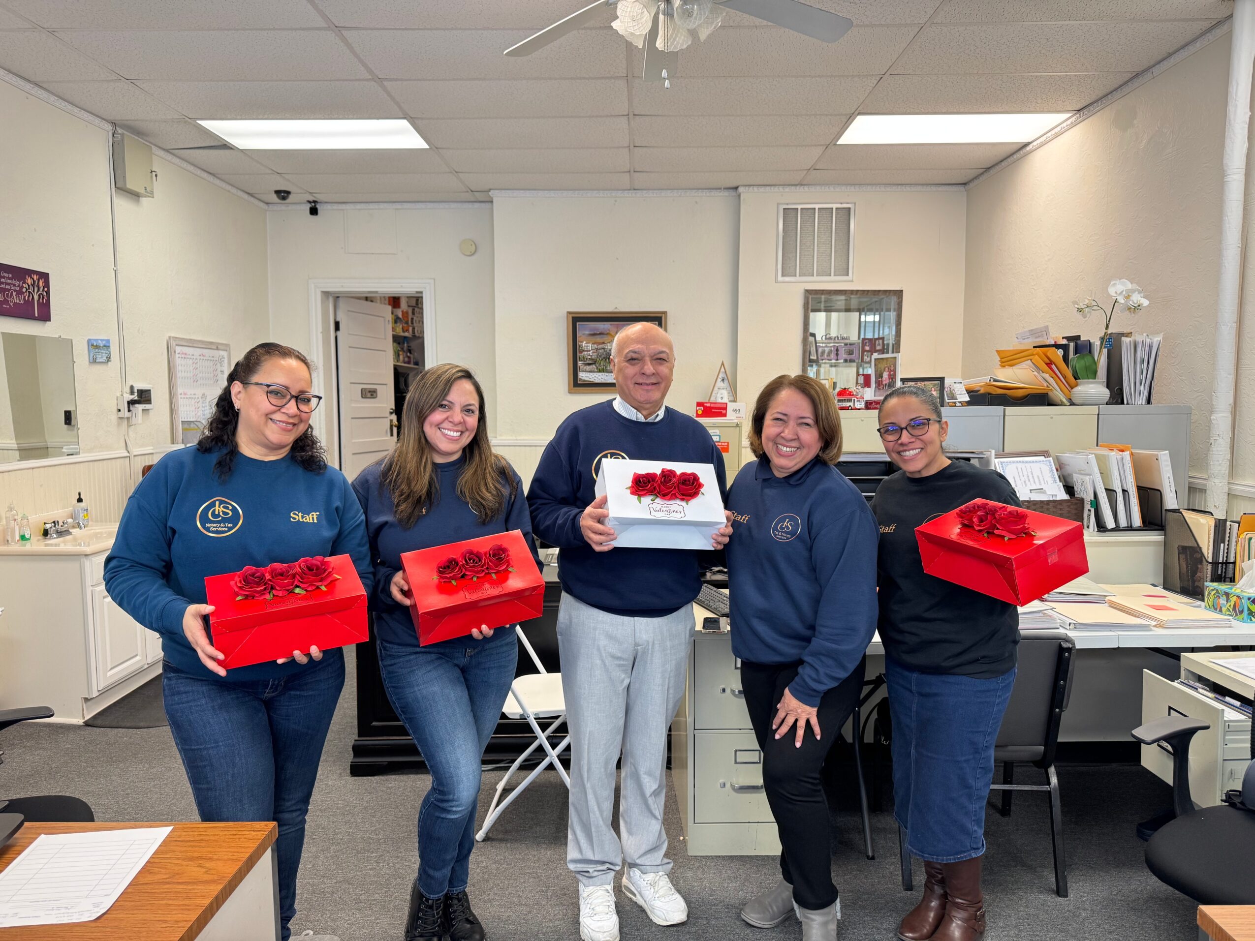 Five people standing in an office holding red boxes, smiling, with office furniture and shelves in the background.