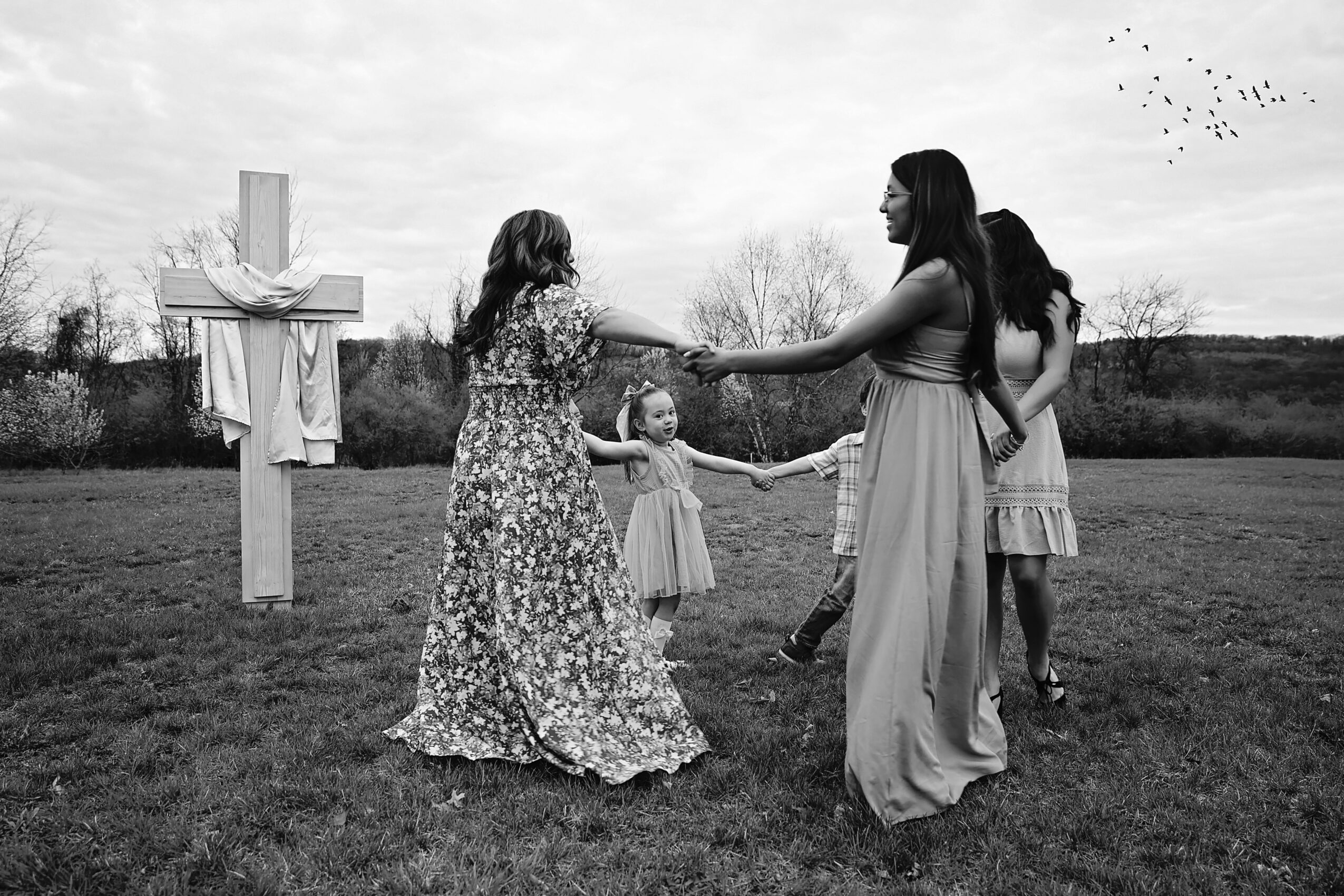 People gather outdoors near a cross, holding hands, with trees and a cloudy sky in the background.