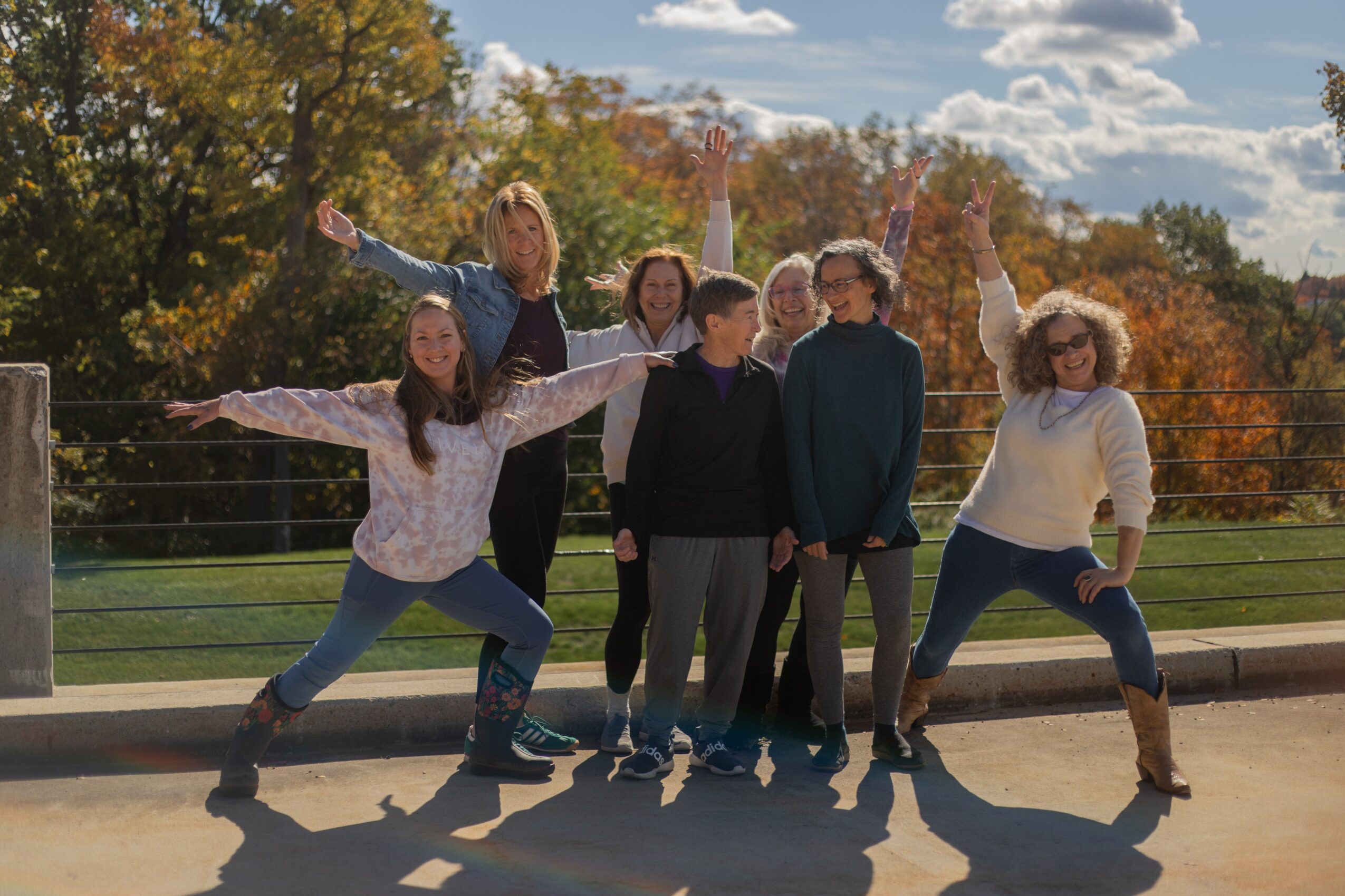 Group of six people outdoors, some jumping and smiling, with trees and sky in background.