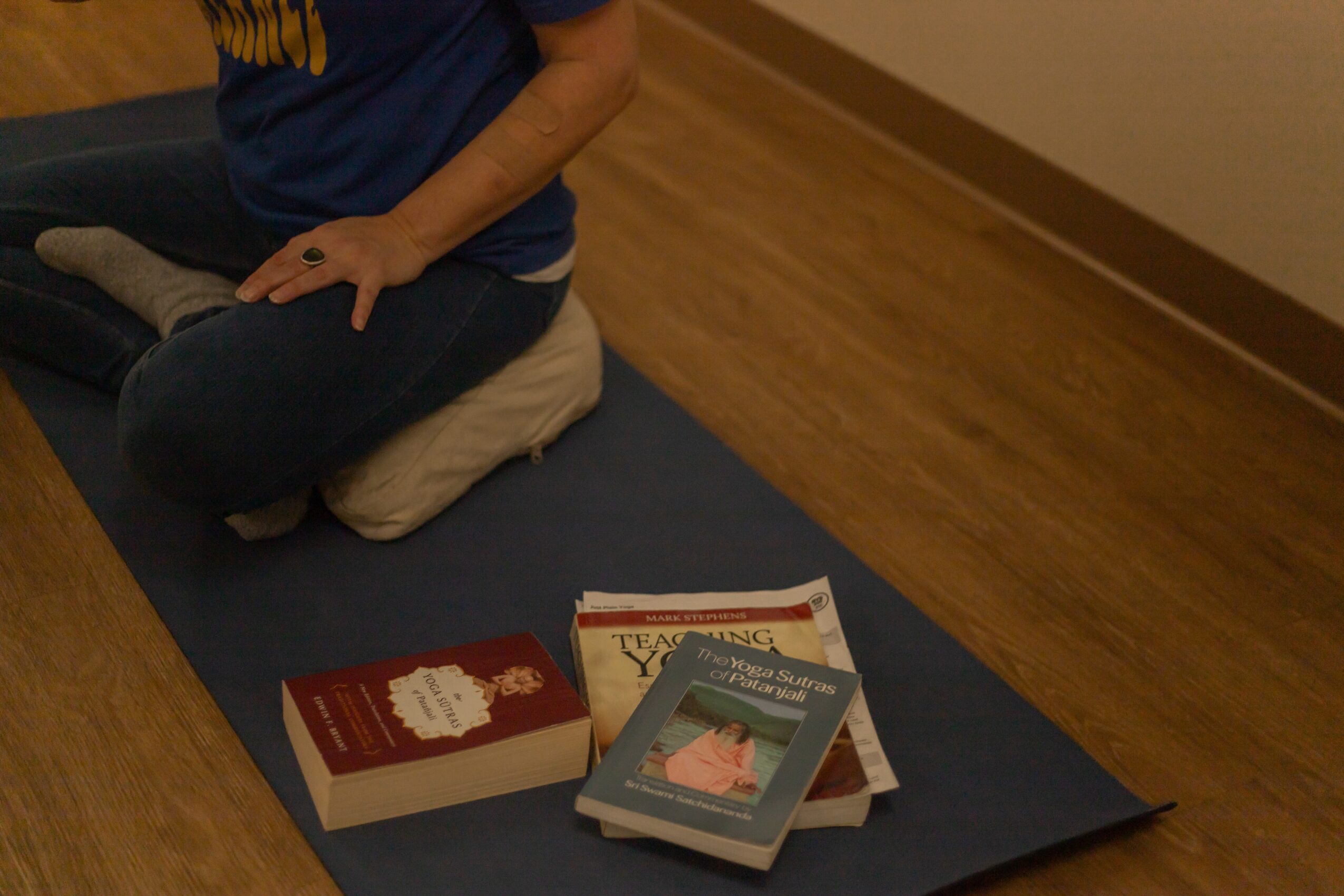 Person sitting cross-legged on a cushion on a yoga mat, with books nearby on a wooden floor.