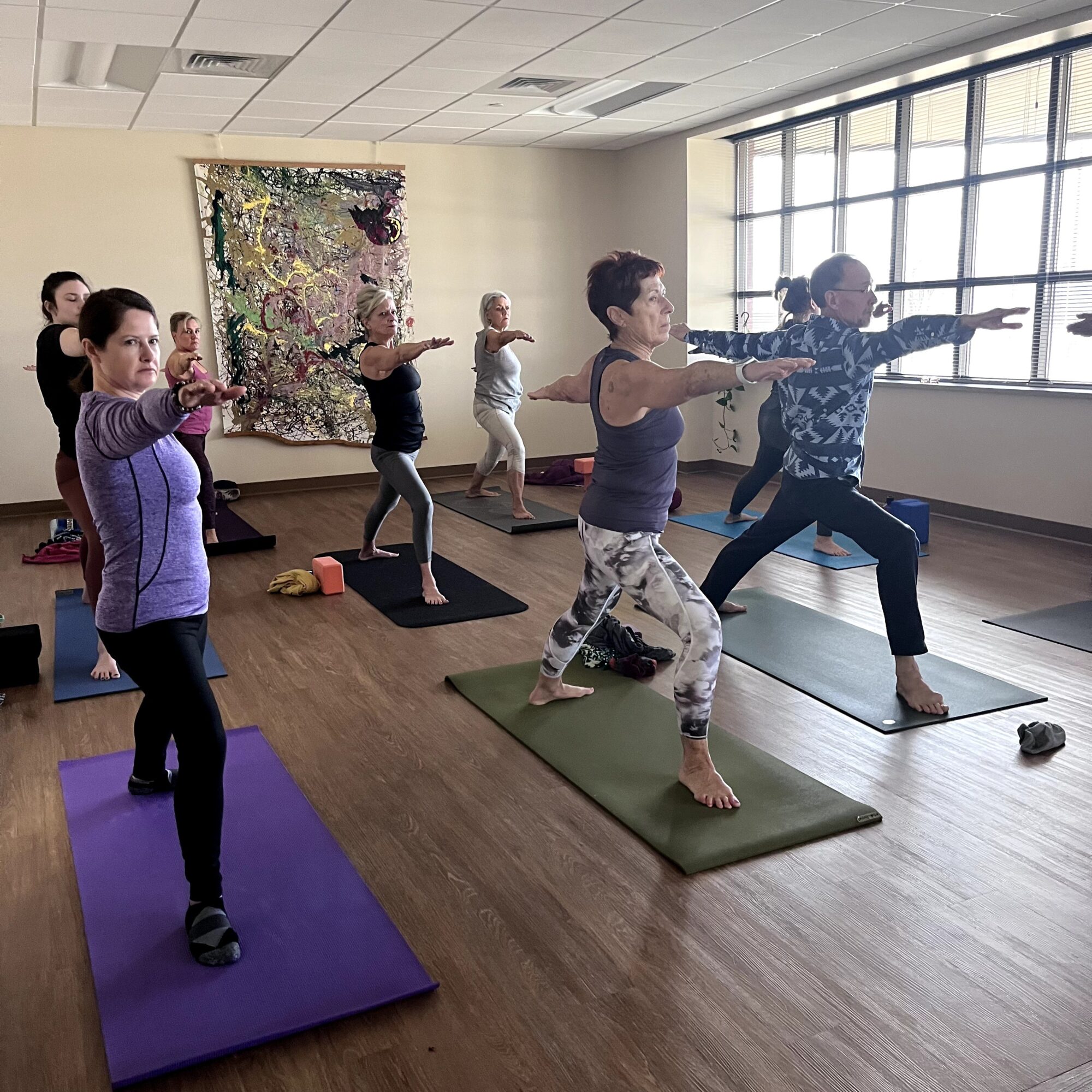 Group of people practicing yoga in a bright room with large windows, standing on mats with arms extended forward.