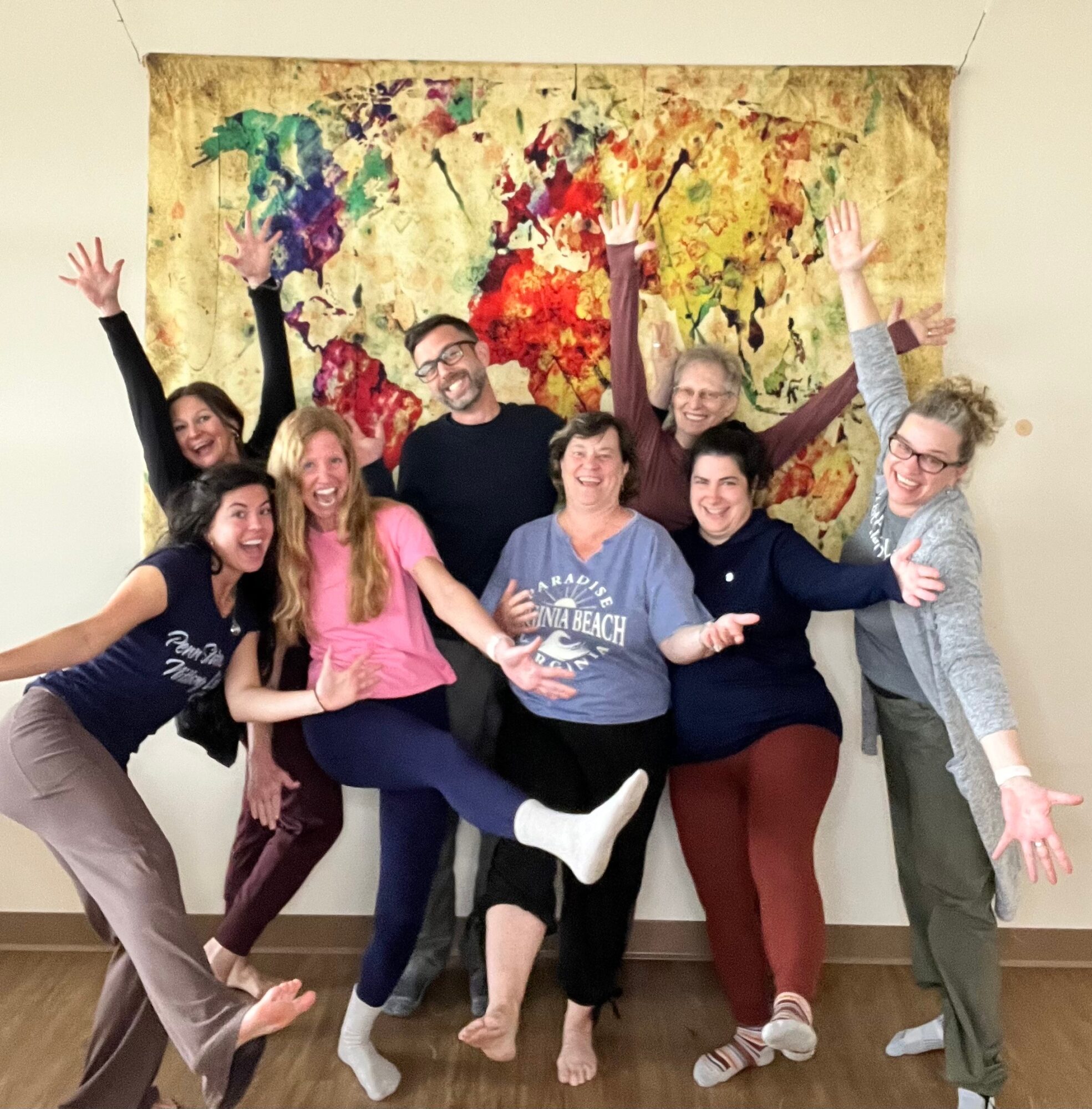 Group of seven people smiling and raising hands in front of a colorful tree painting, standing indoors.