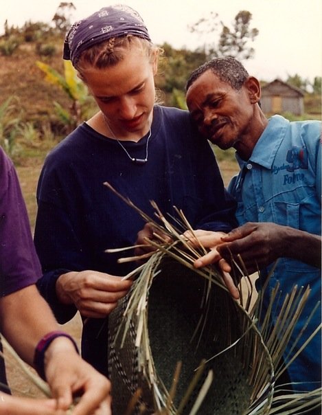 Two people, one young woman and one man, look at a plant or crop outdoors with greenery in the background.
