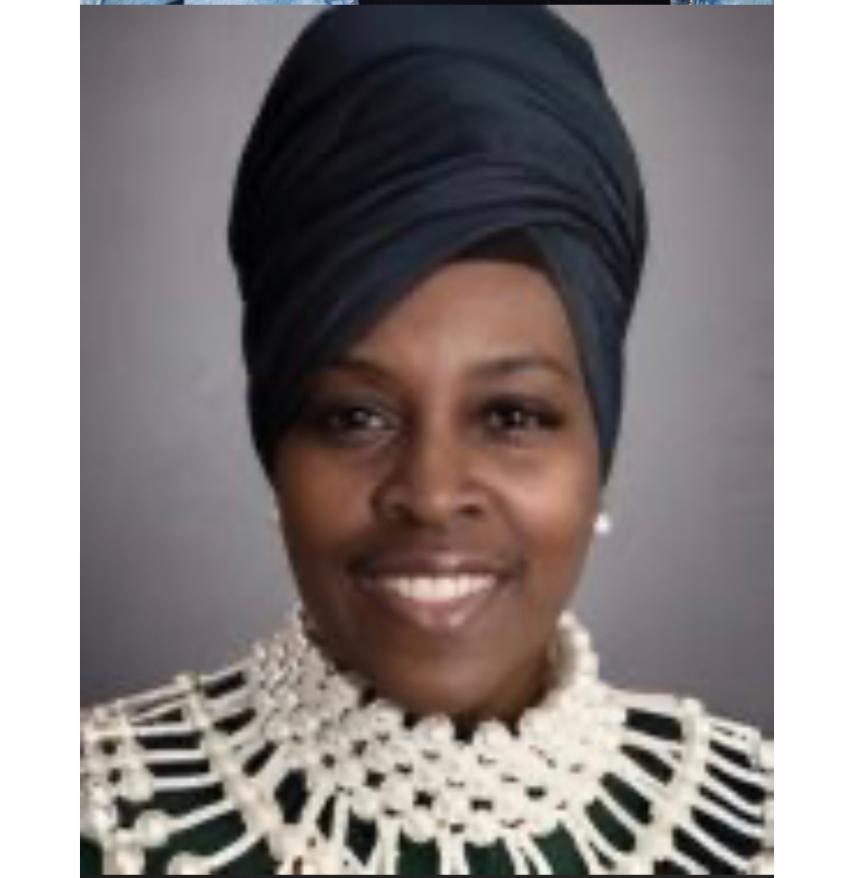 Portrait of a woman wearing a headwrap and a large pearl necklace, smiling, against a plain background.