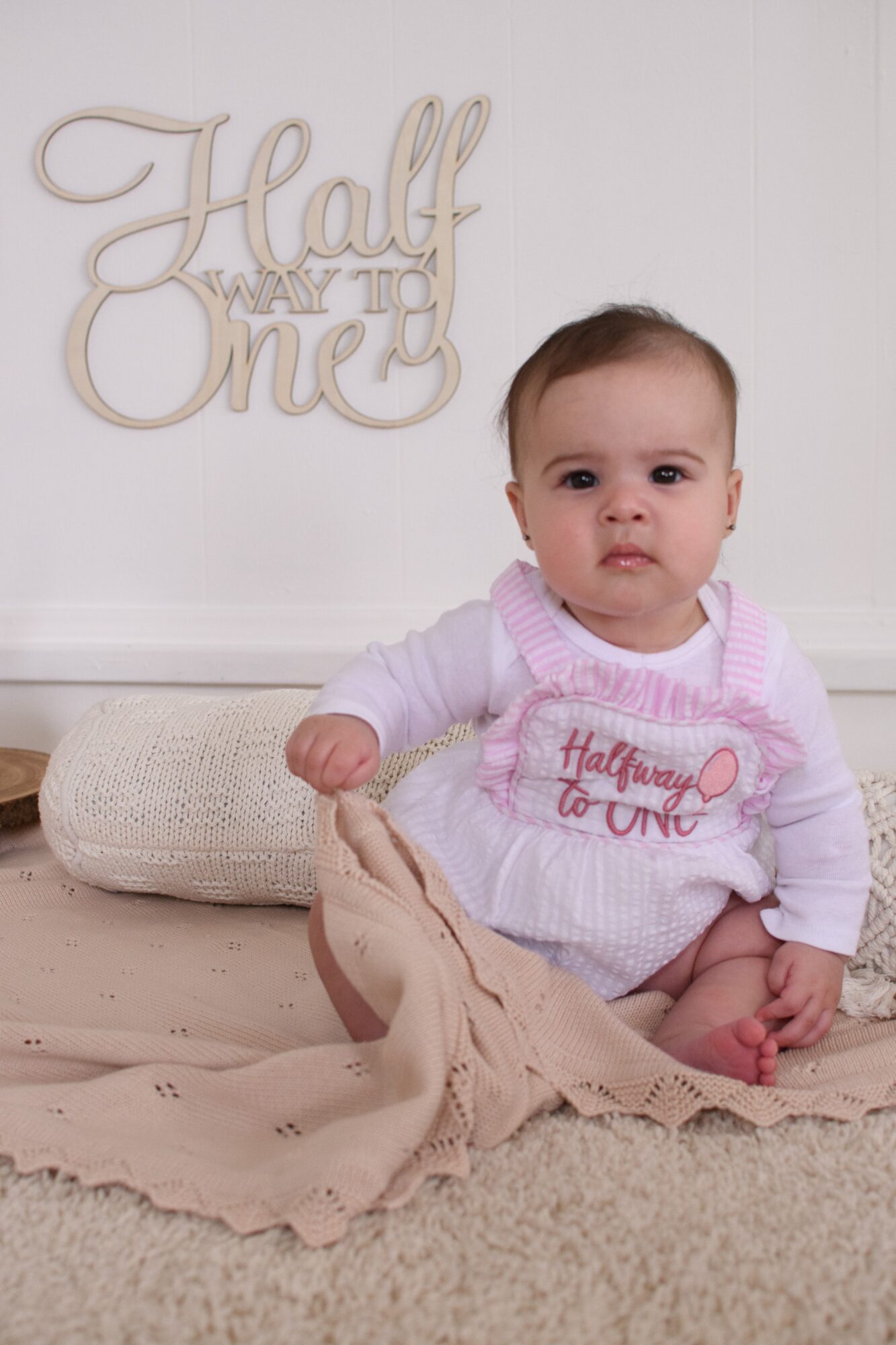 Baby girl sitting on carpet with beige blanket, wearing a pink and white outfit, in front of a wall with a decorative sign.