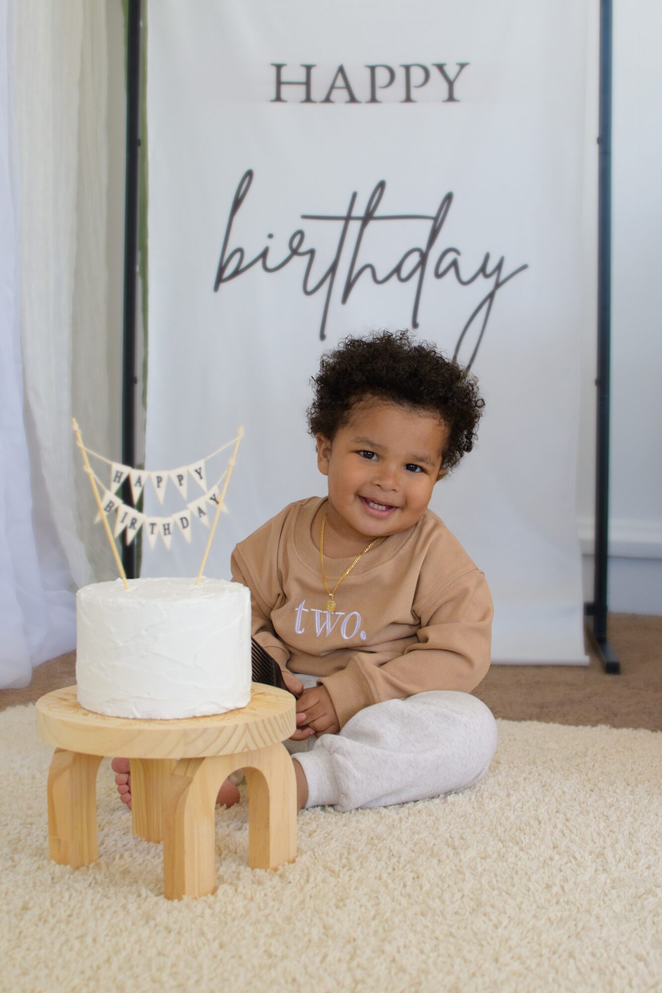 Child sitting on floor next to a birthday cake with a banner, smiling, in front of a 'Happy birthday' sign.