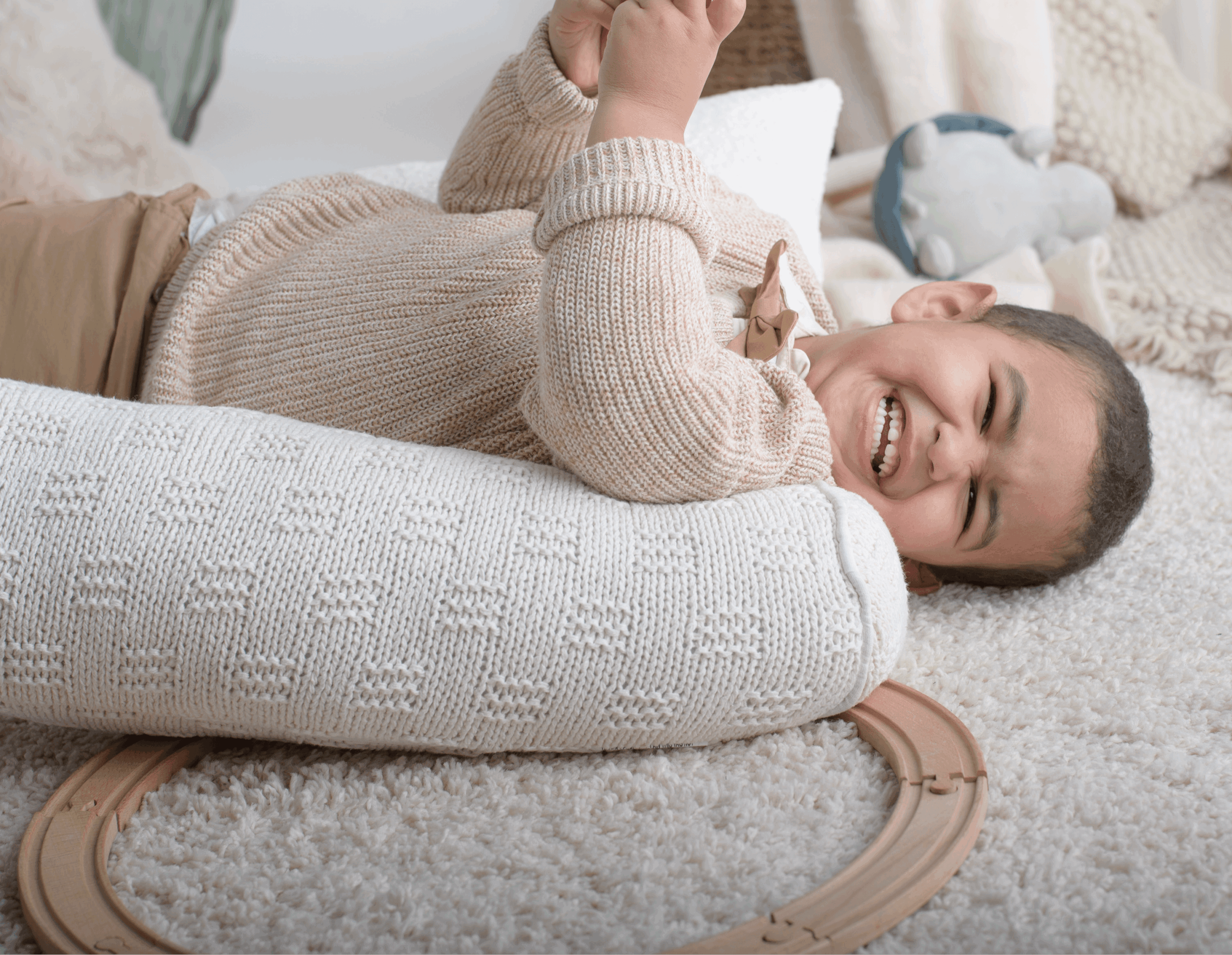 Child lying on a carpet, smiling and holding a toy, with a train track and plush toys nearby.