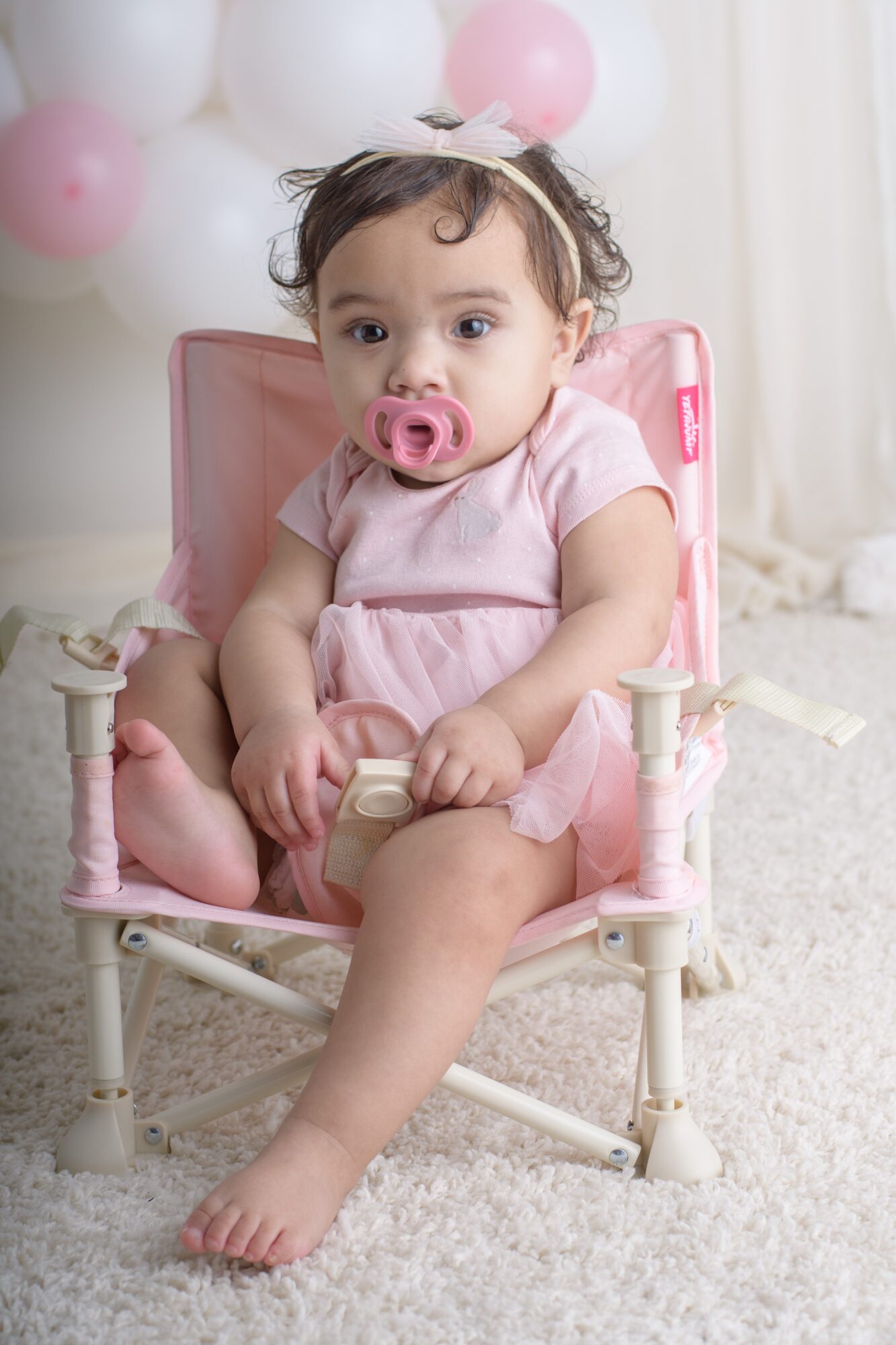 Young girl with a pacifier sitting in a pink chair, surrounded by pink and white balloons, on a white carpet.