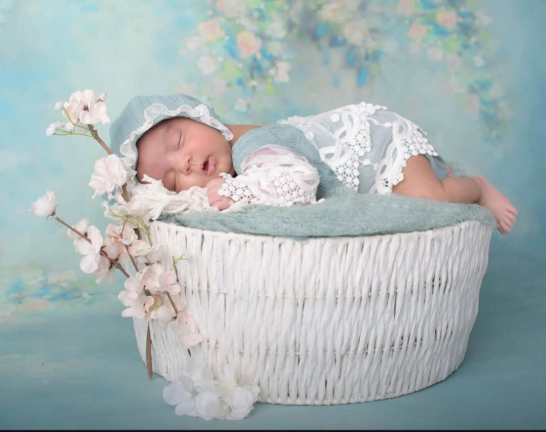 Sleeping baby in a lace outfit lying on a soft cushion inside a white basket, with flowers nearby, against a pastel background.