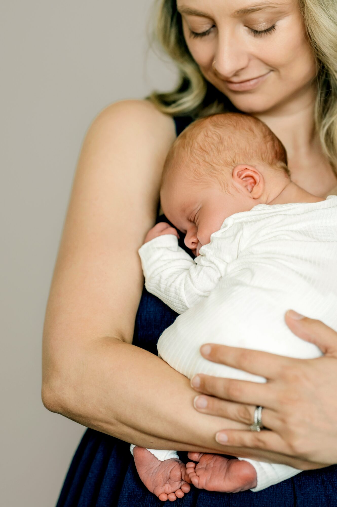Woman holding a sleeping baby close to her chest, smiling gently, with a neutral background.