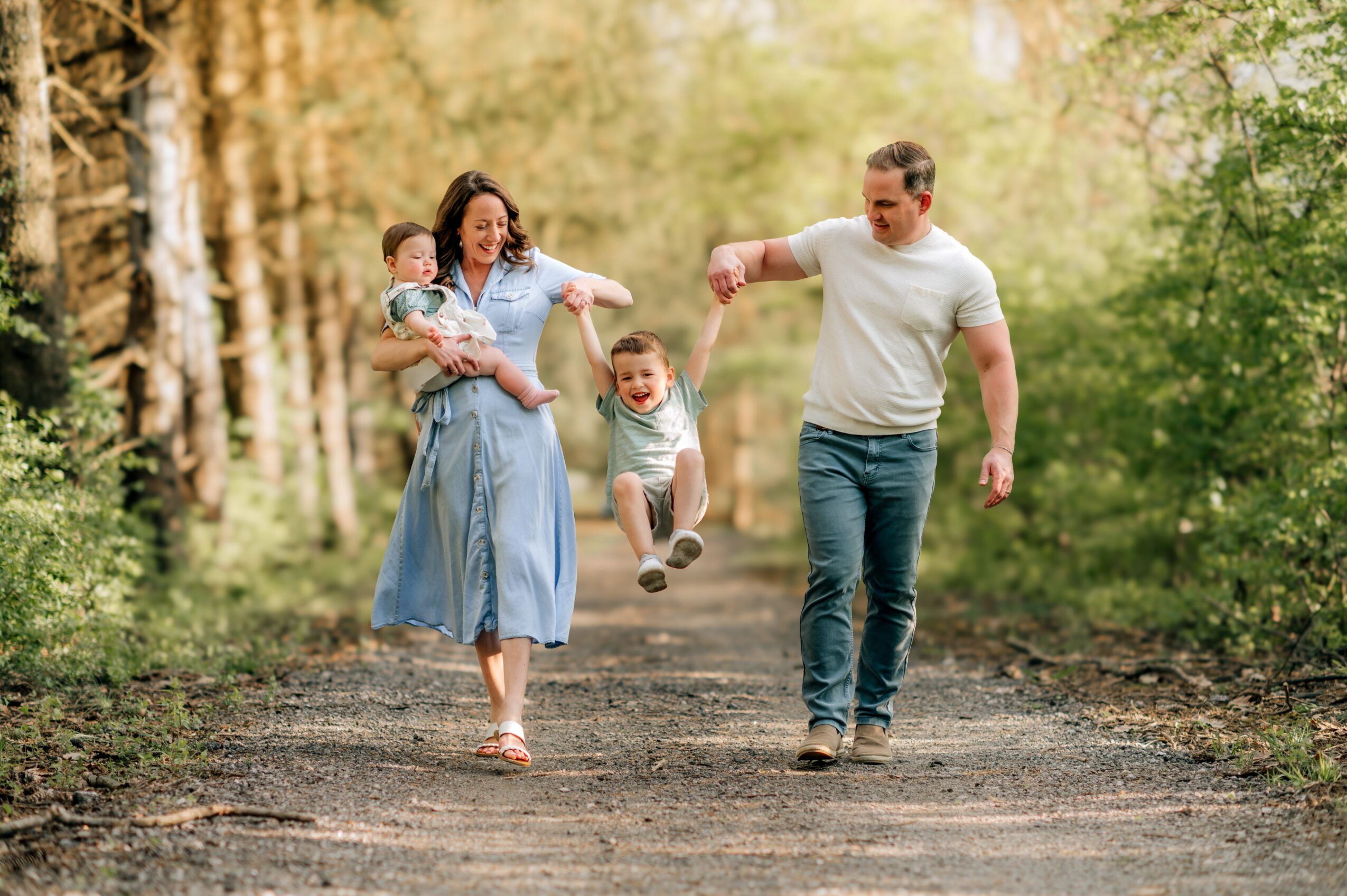 Family walking and playing in a forest, children jumping, adults smiling, trees and sunlight in background.