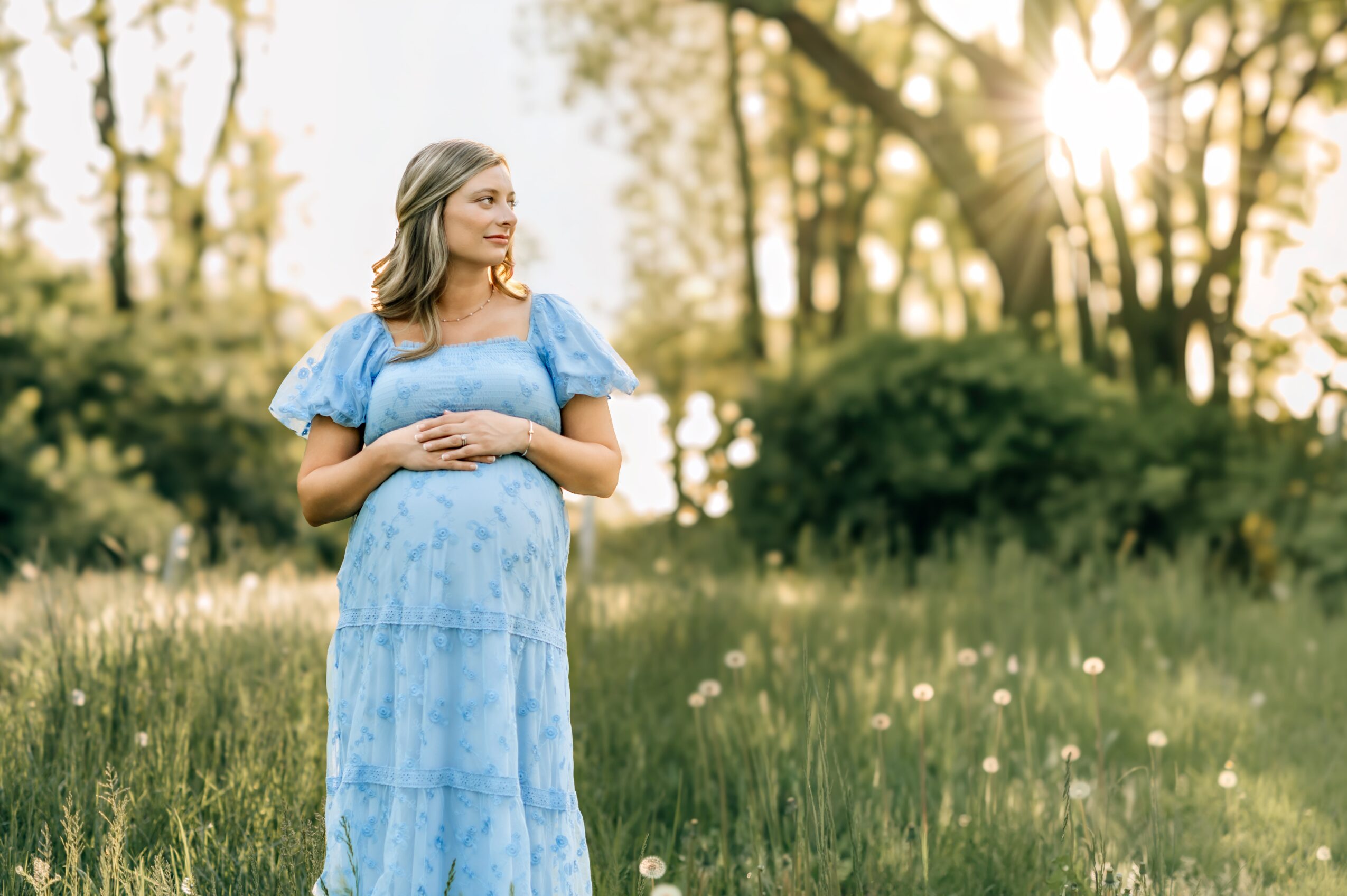 Pregnant woman in a blue dress standing in a grassy field with trees and sunlight in the background.