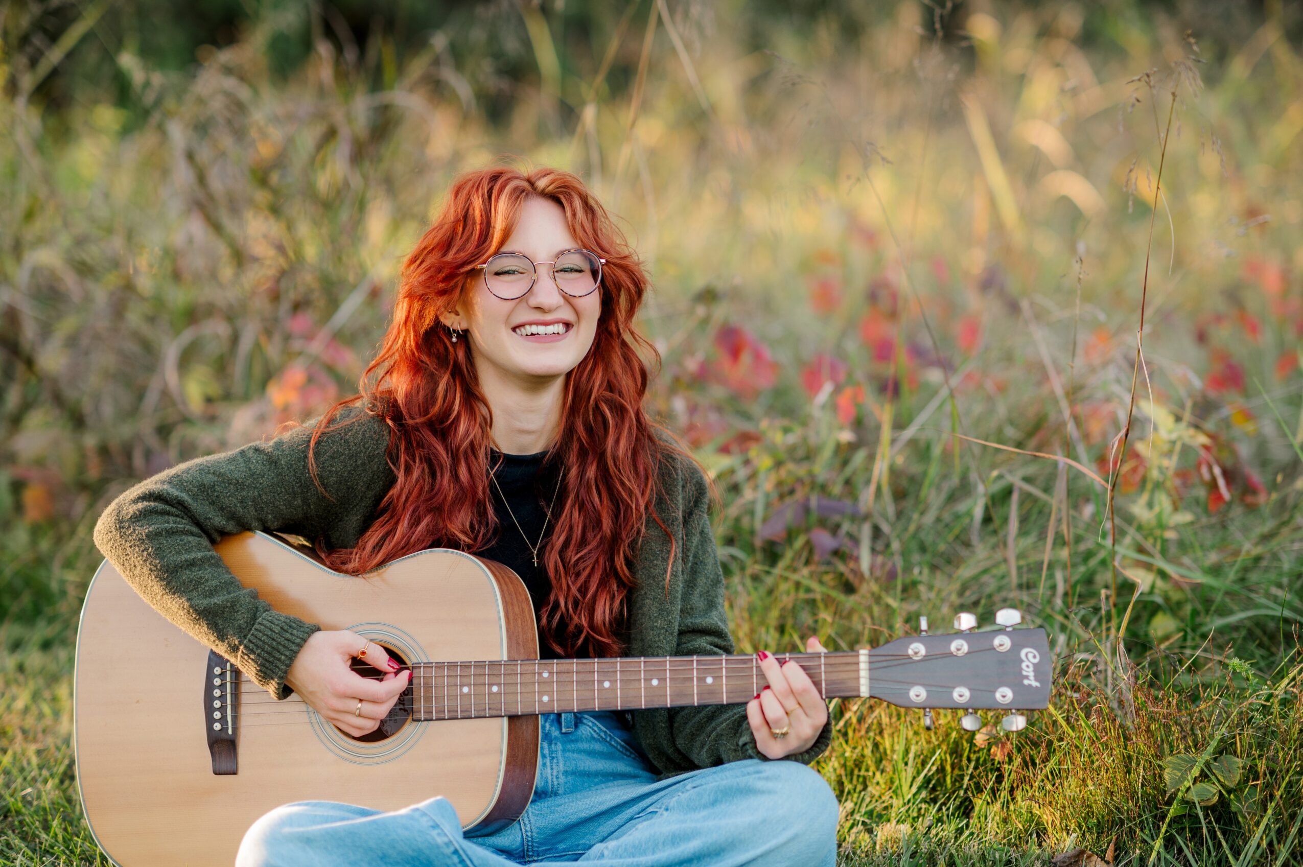 Young woman with red hair and glasses playing guitar outdoors in a grassy field.