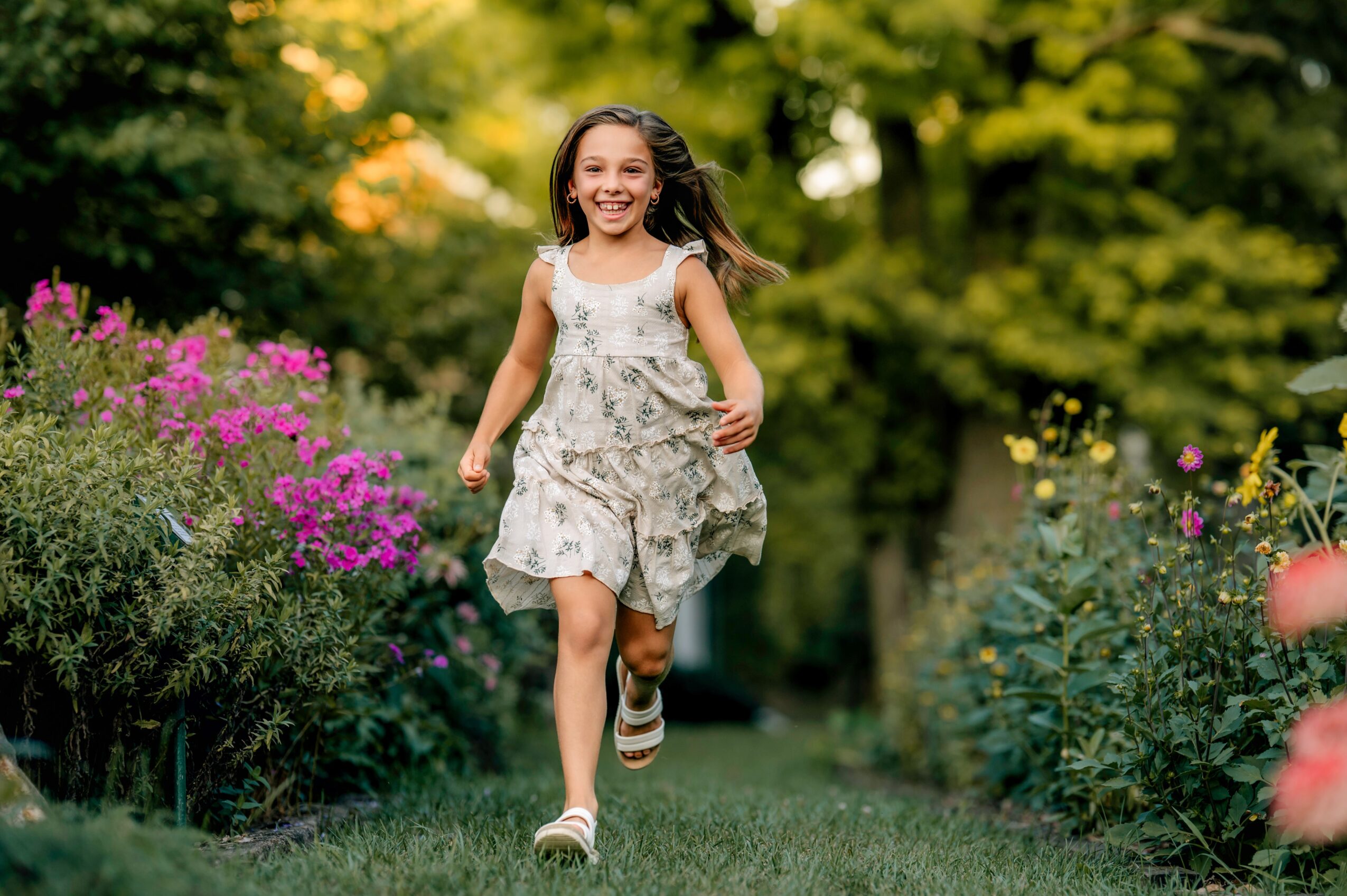 Young girl running through a garden with colorful flowers and green trees, smiling happily.