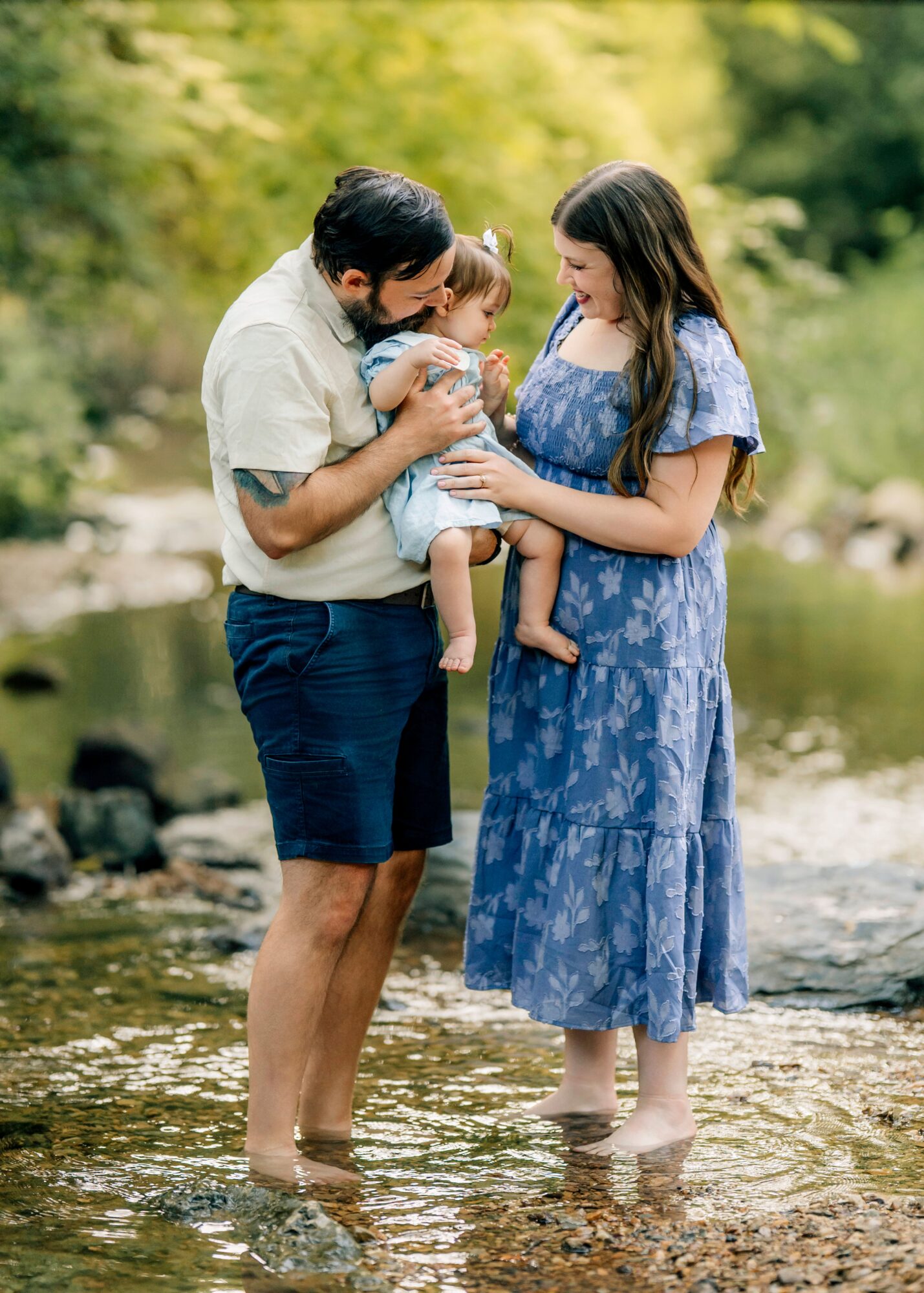 Family of three standing in a shallow river, holding a baby, surrounded by greenery, smiling at each other.