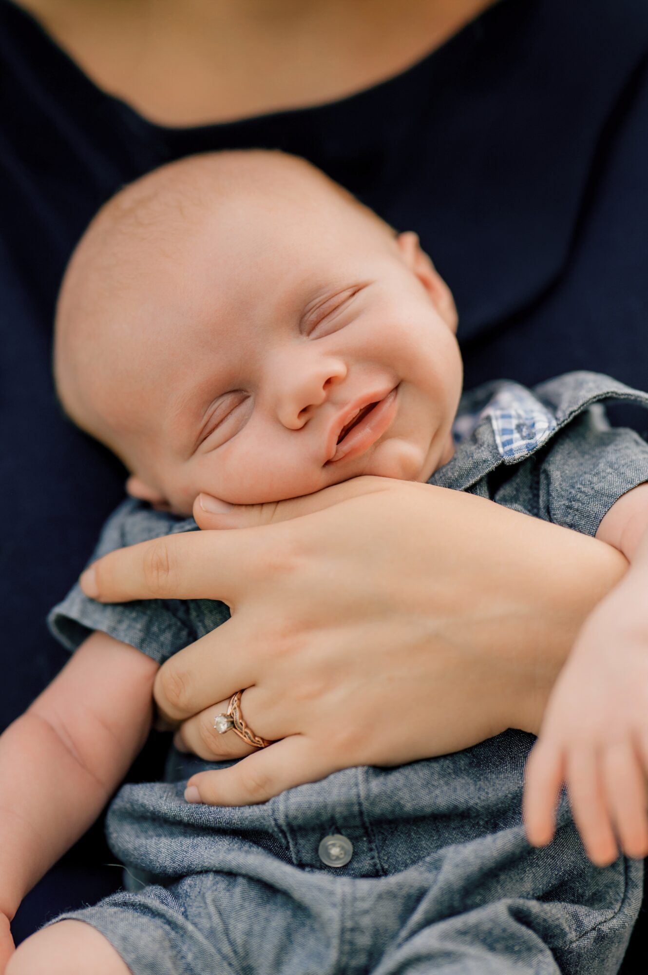 Baby with closed eyes smiling, being held gently by a person, wearing a denim outfit, in a close-up shot.