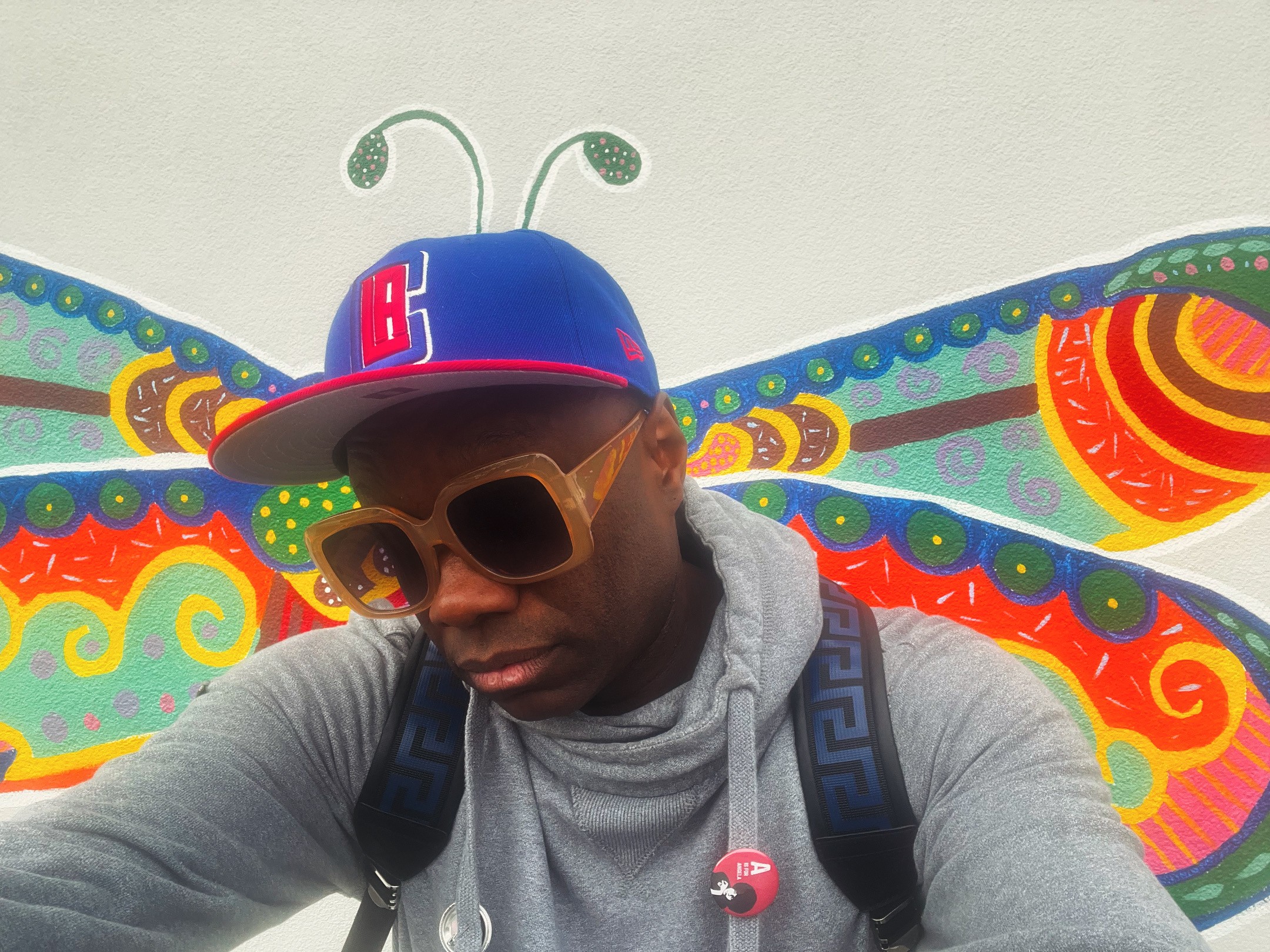 Person wearing sunglasses and a cap with antennae, standing in front of colorful butterfly wings backdrop.