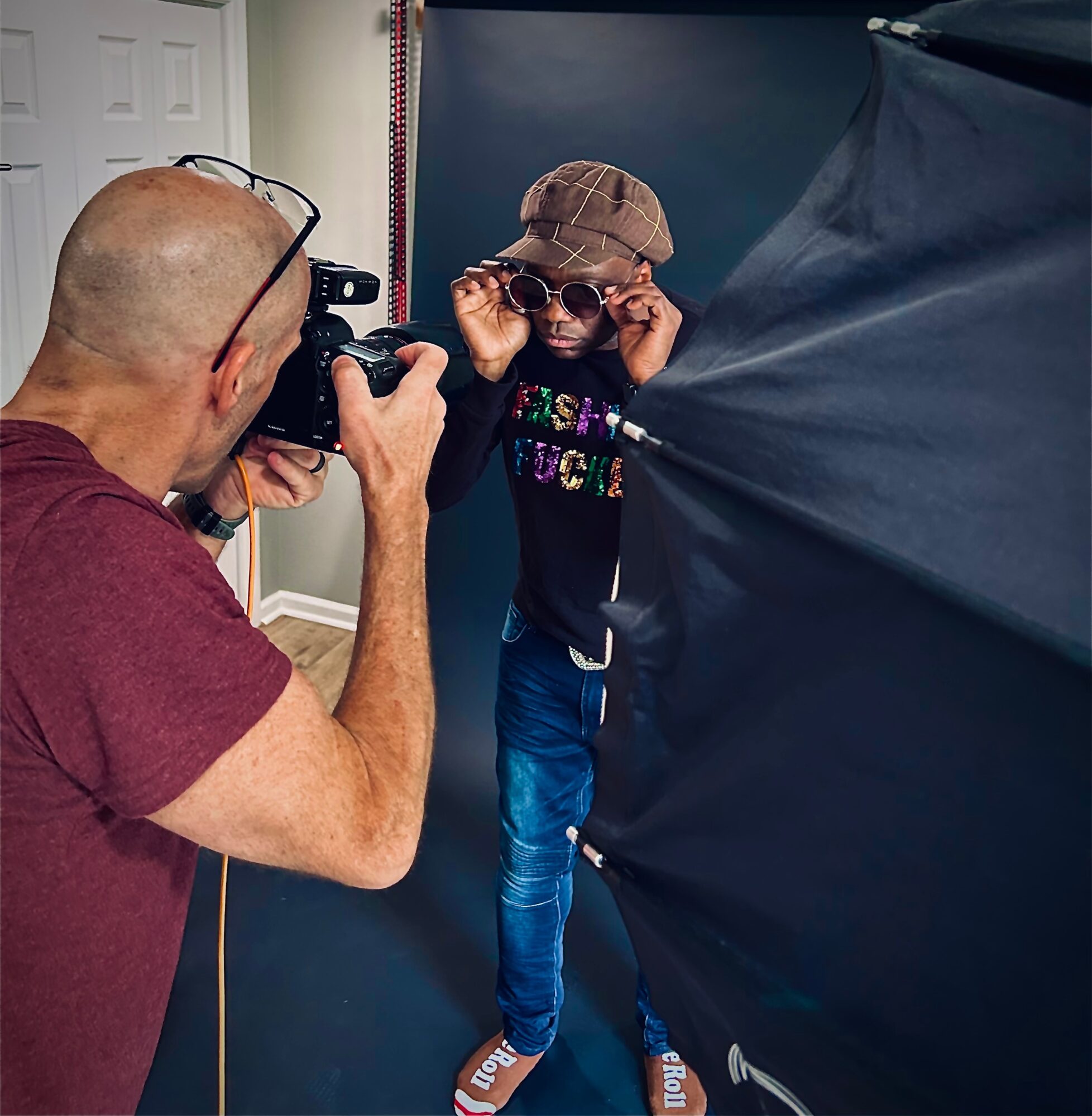 Child wearing sunglasses and a hat poses for a photo, photographer taking picture indoors, dark backdrop, child adjusting sunglasses.