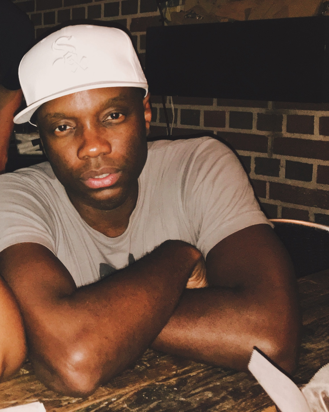 Man wearing a white cap and light-colored shirt sitting at a table with arms crossed, brick wall background.