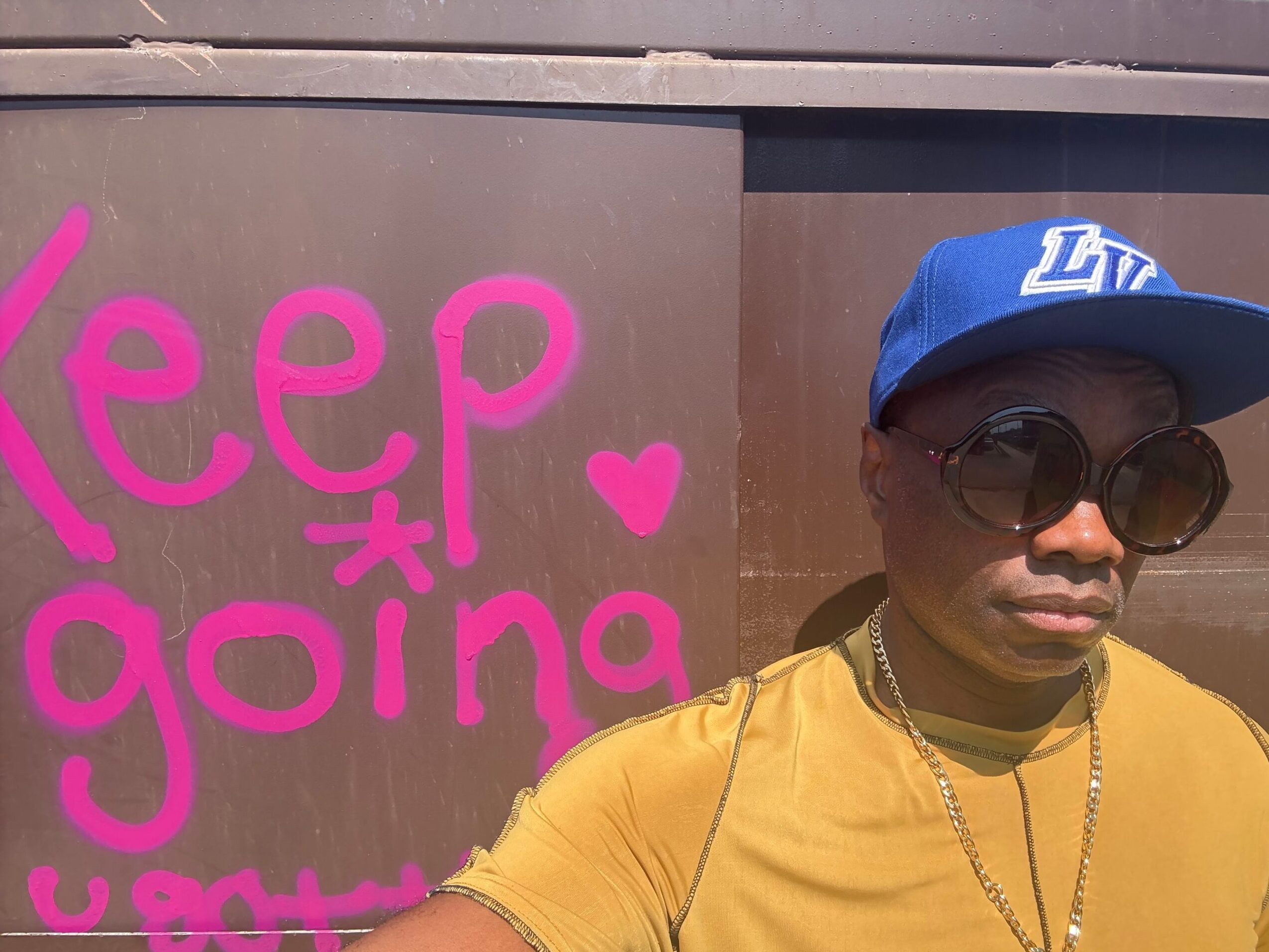 Person wearing sunglasses, a blue cap, and a yellow shirt, standing next to pink writing on a wall.