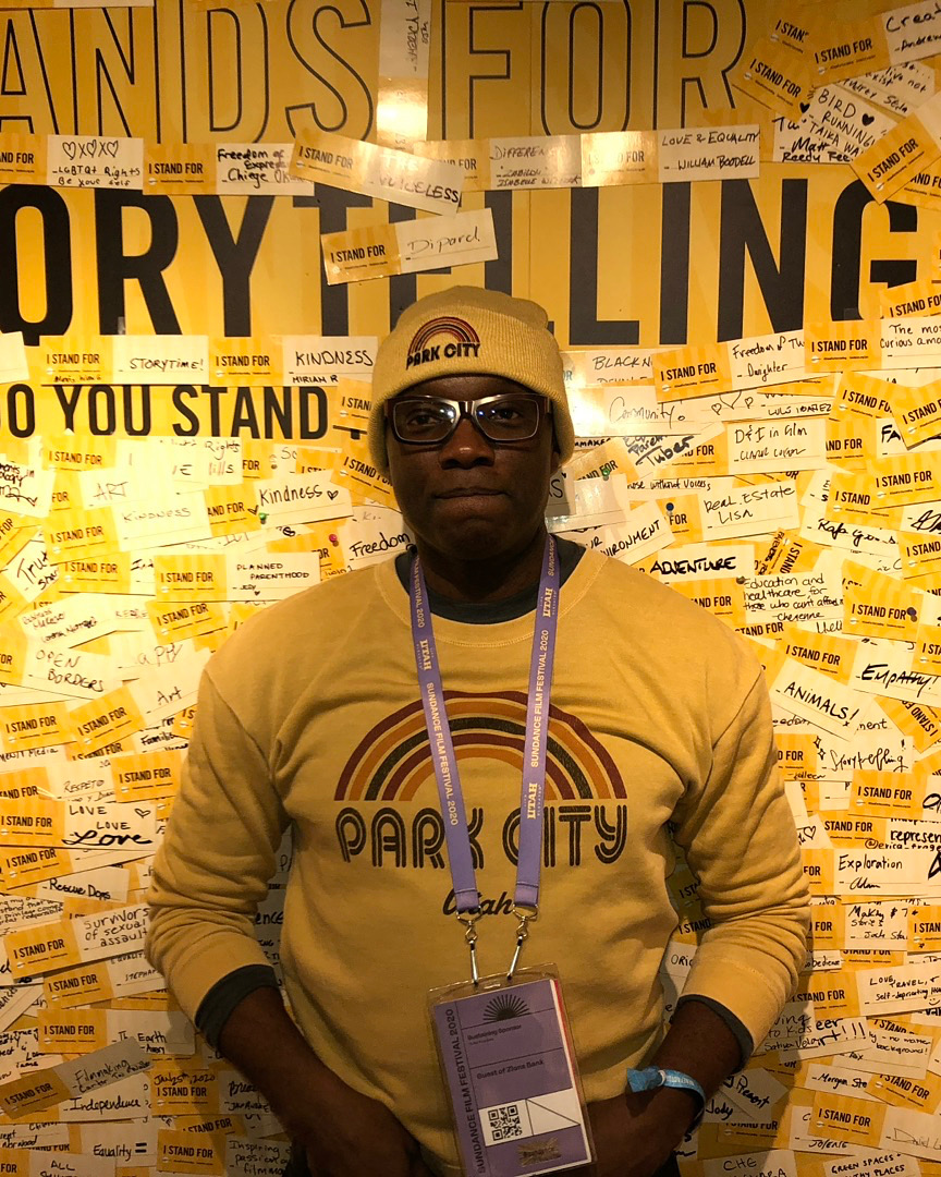 Person wearing glasses and a yellow beanie, standing in front of a yellow background with protest signs and messages.