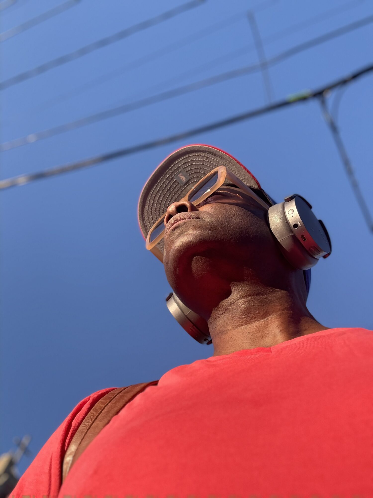 Person wearing sunglasses, headphones, and a red shirt, looking upward against a blue sky with power lines.