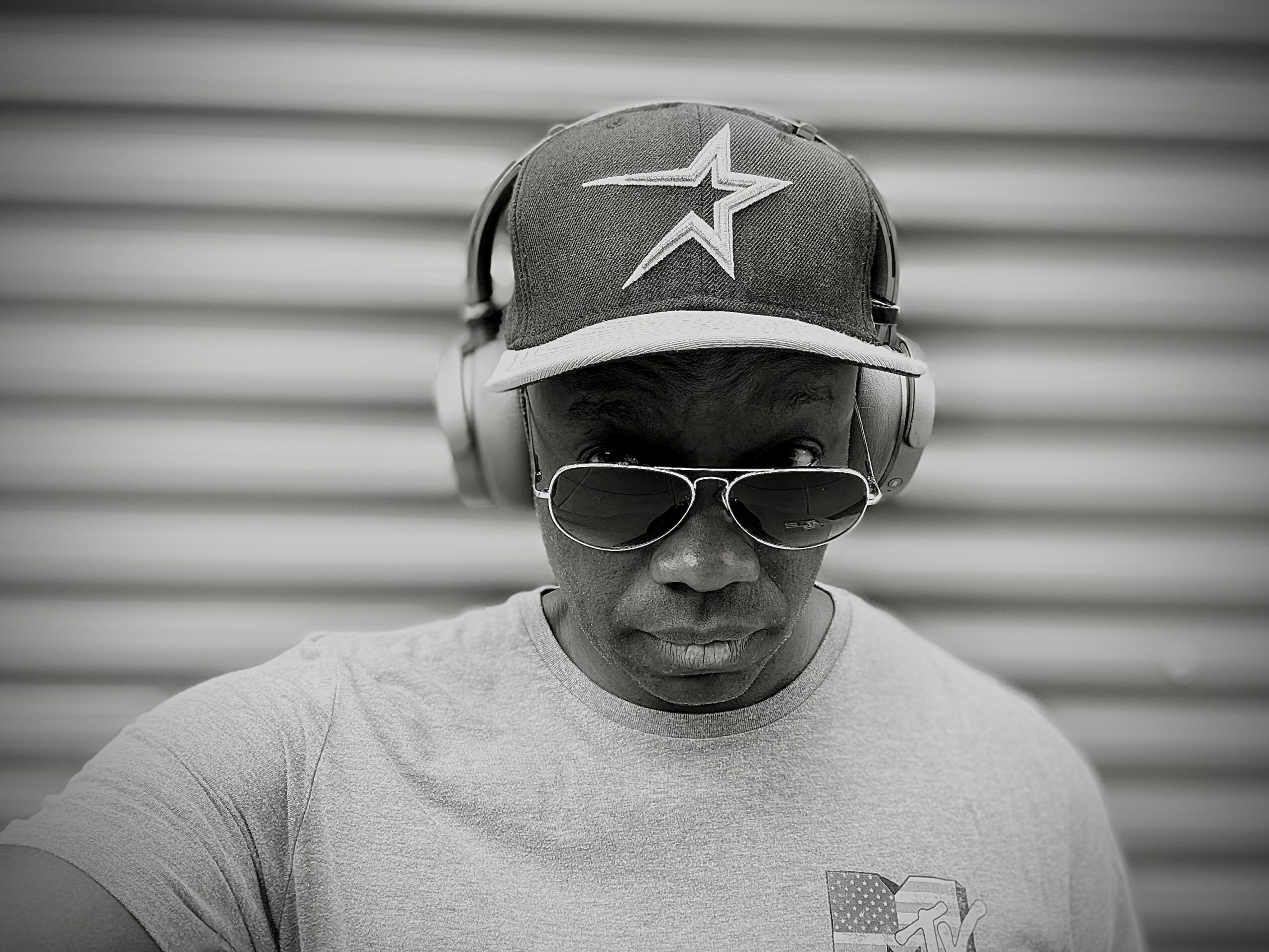 Person wearing sunglasses, headphones, and a cap with a star logo, looking directly at the camera, background with horizontal lines.
