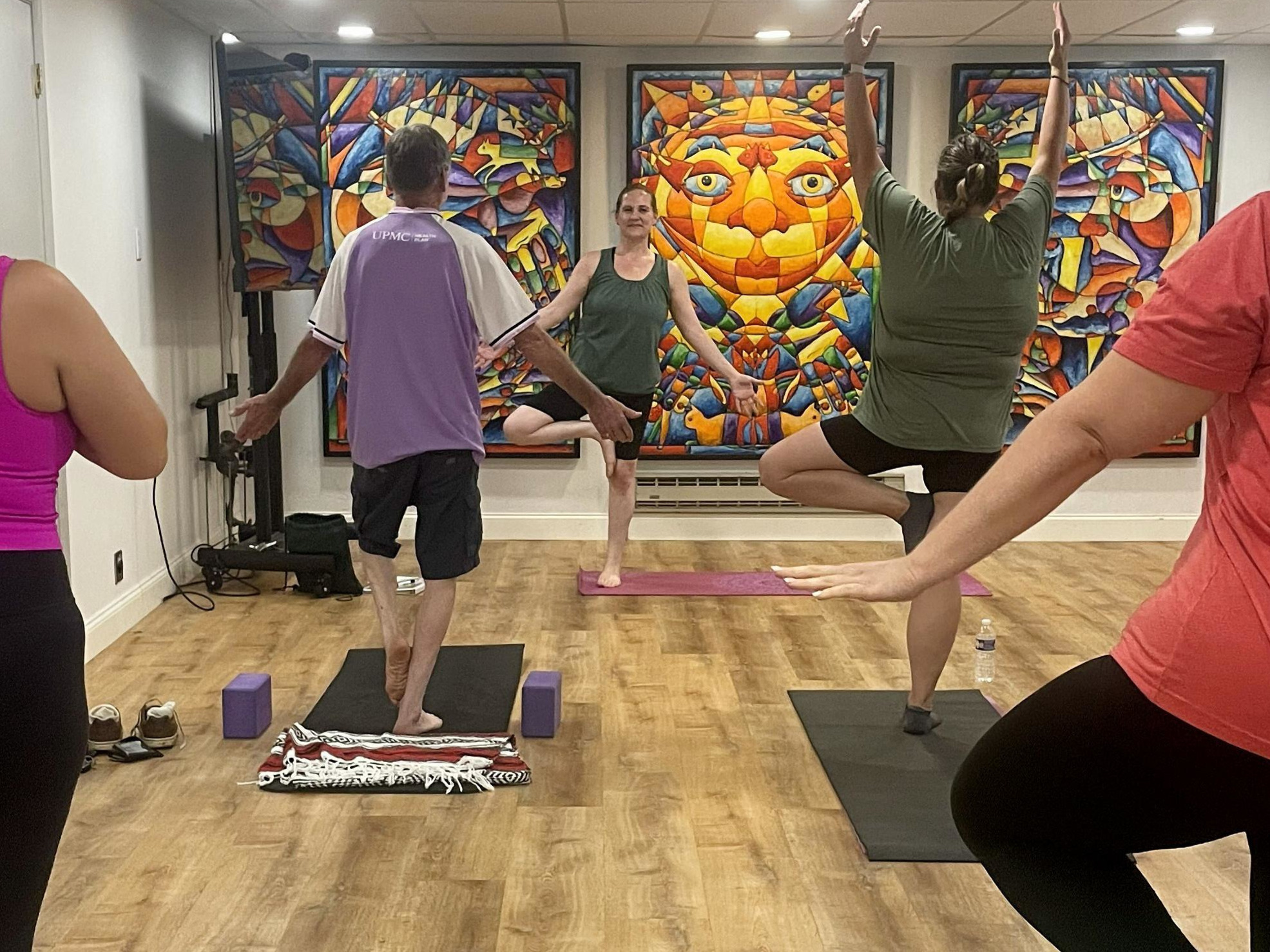 Group of people practicing yoga in a studio with colorful abstract paintings on the wall, some standing on mats, one in a tree pose.