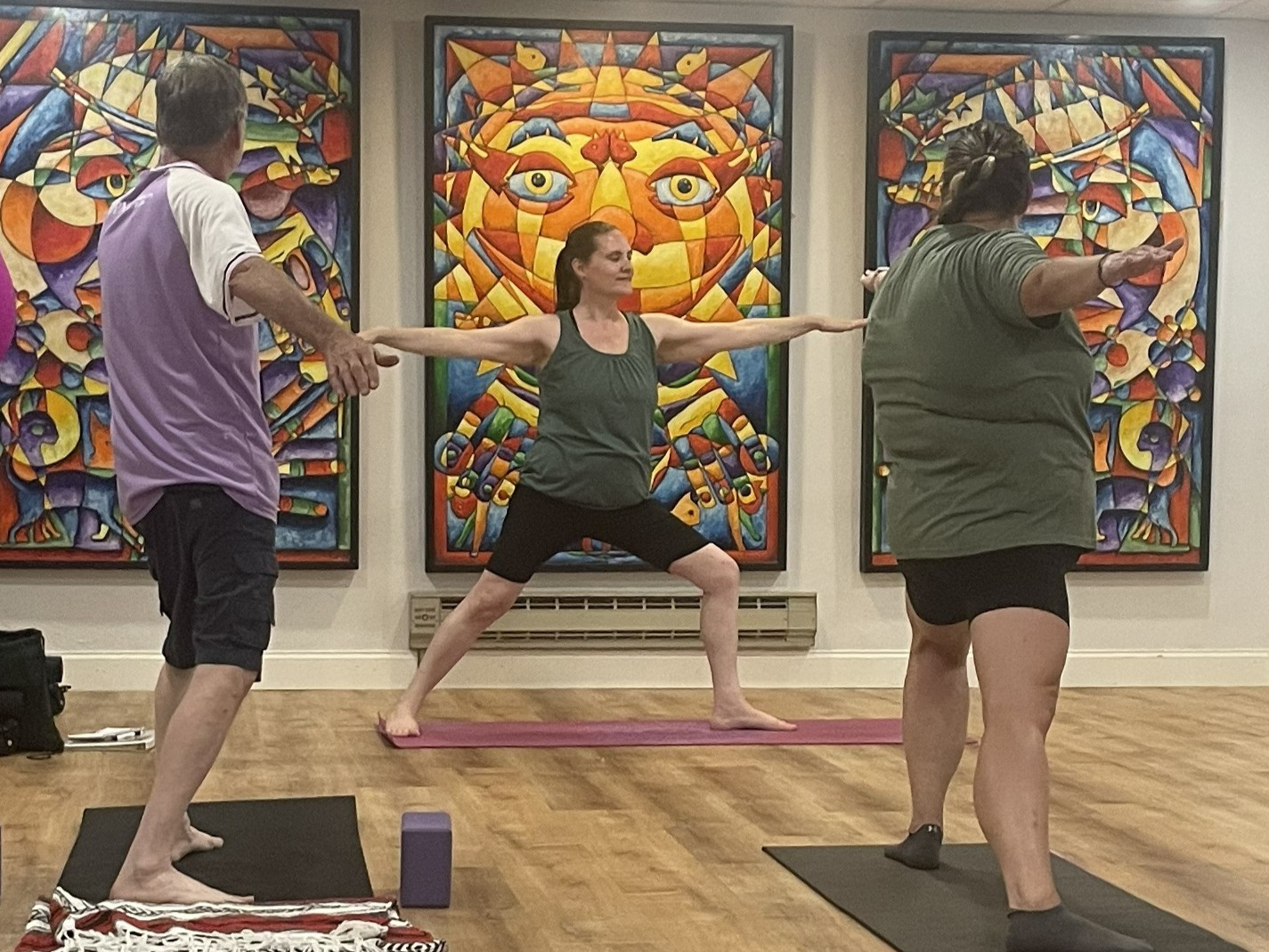 Three people practicing yoga in an art gallery with colorful paintings, one person in a warrior pose, two others holding hands.