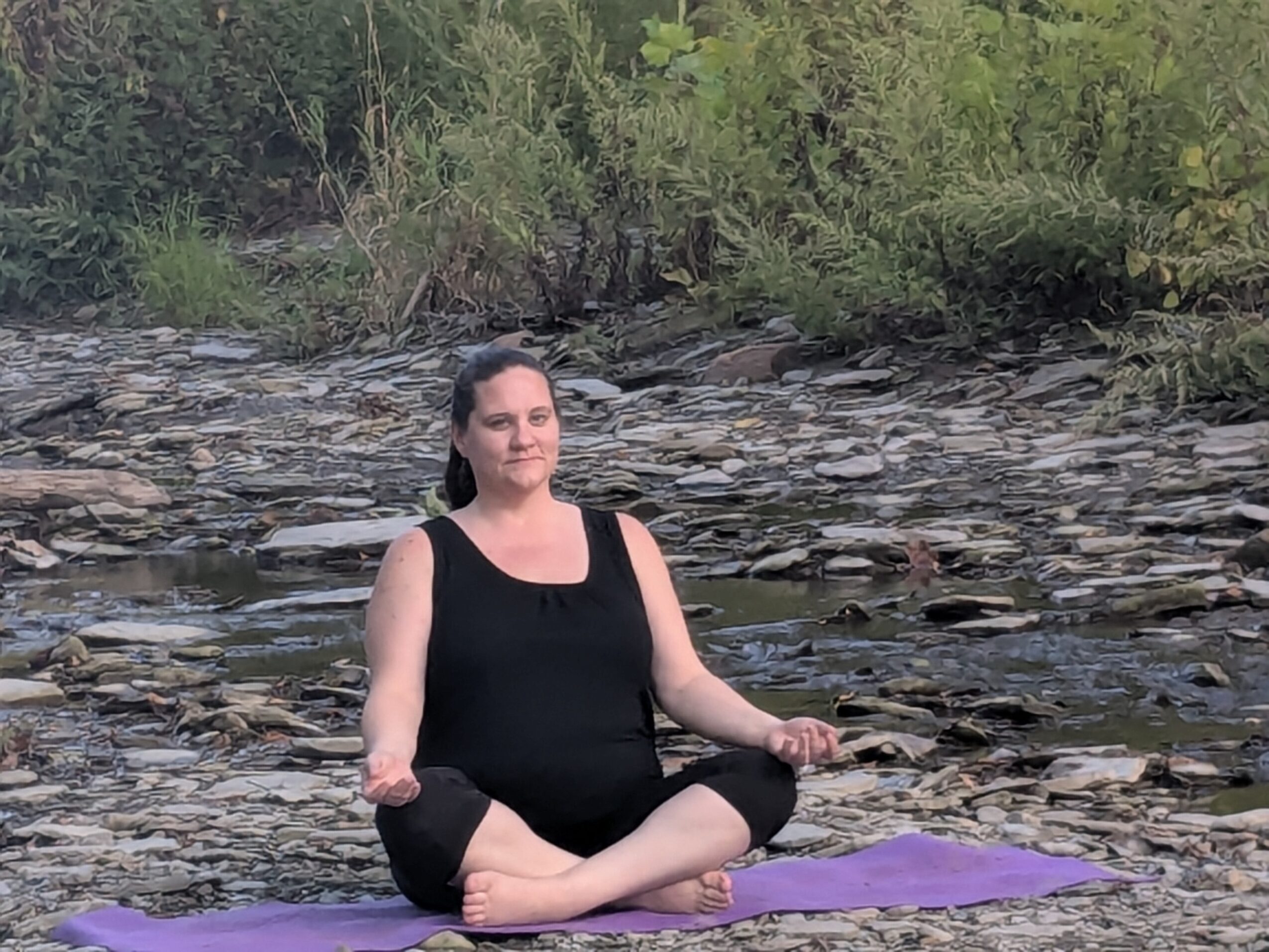 Woman practicing yoga outdoors on a purple mat near a rocky riverbank, surrounded by greenery.