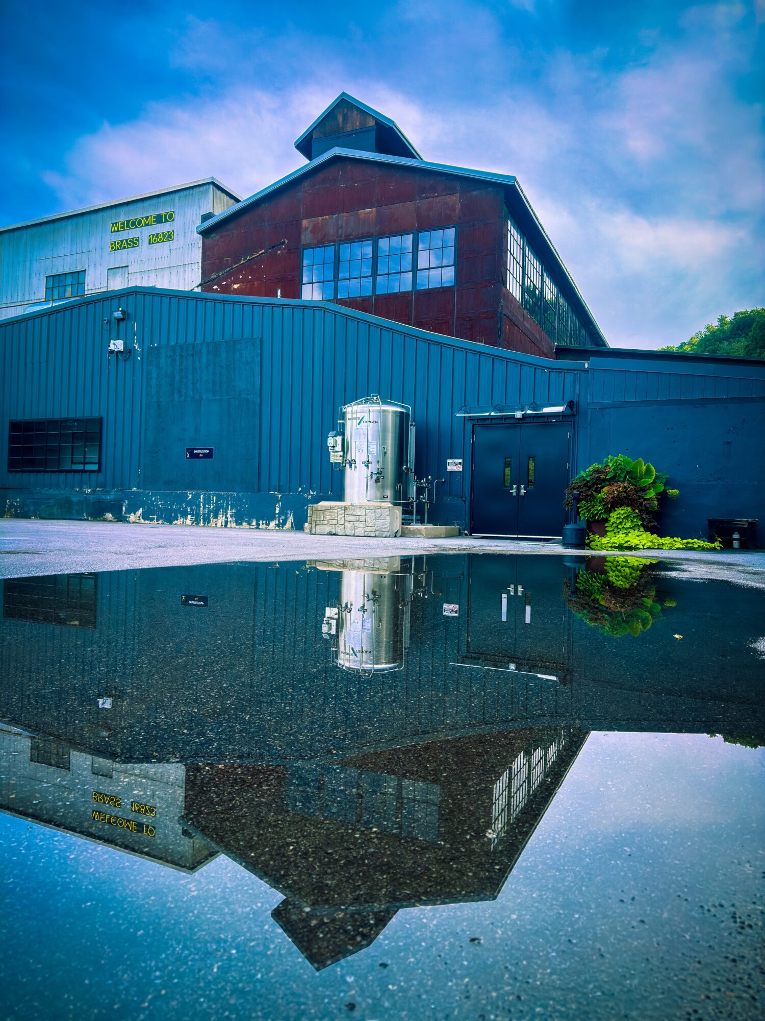 Building with blue and red sections reflected in a puddle, sky with clouds above, and greenery nearby.