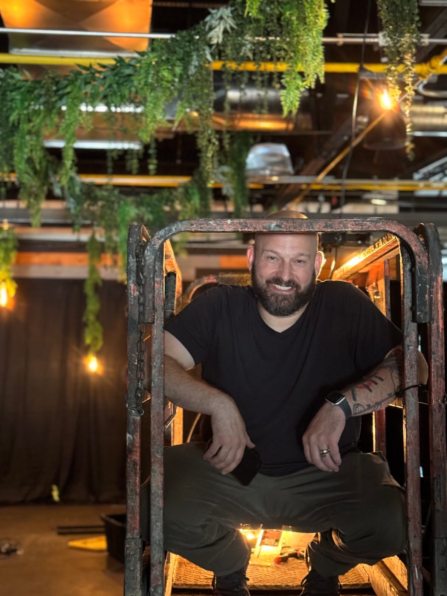 Man with a beard sitting on a chair, smiling, in a decorated indoor space with hanging plants and warm lighting.