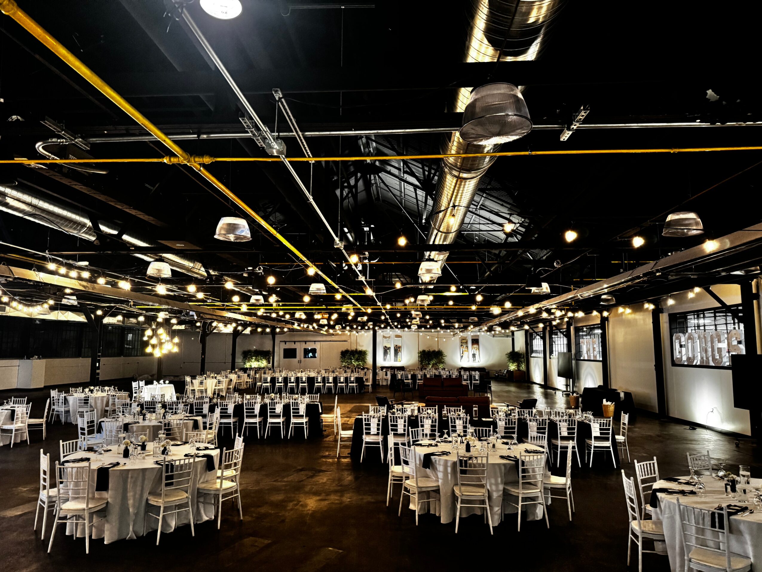 Large event space with round tables and chairs, decorated for a formal gathering, with ceiling lights and industrial ceiling design.