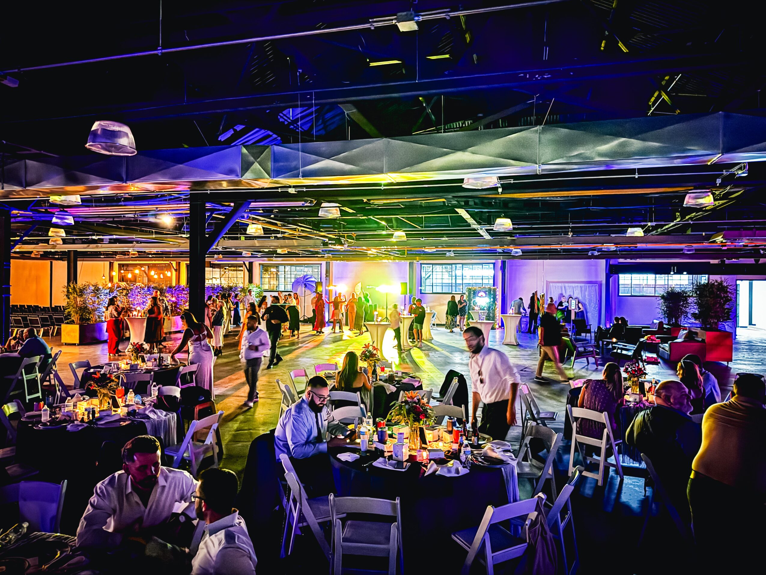 People gather in a brightly lit event space with tables, chairs, and colorful lighting effects.