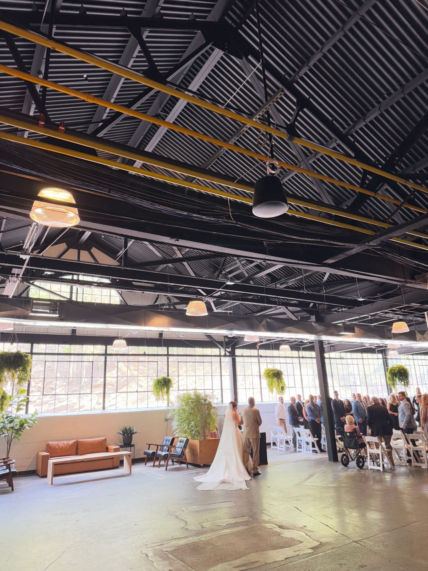 People gather in a spacious indoor area with large windows, tables, and chairs, some standing and talking, others sitting.