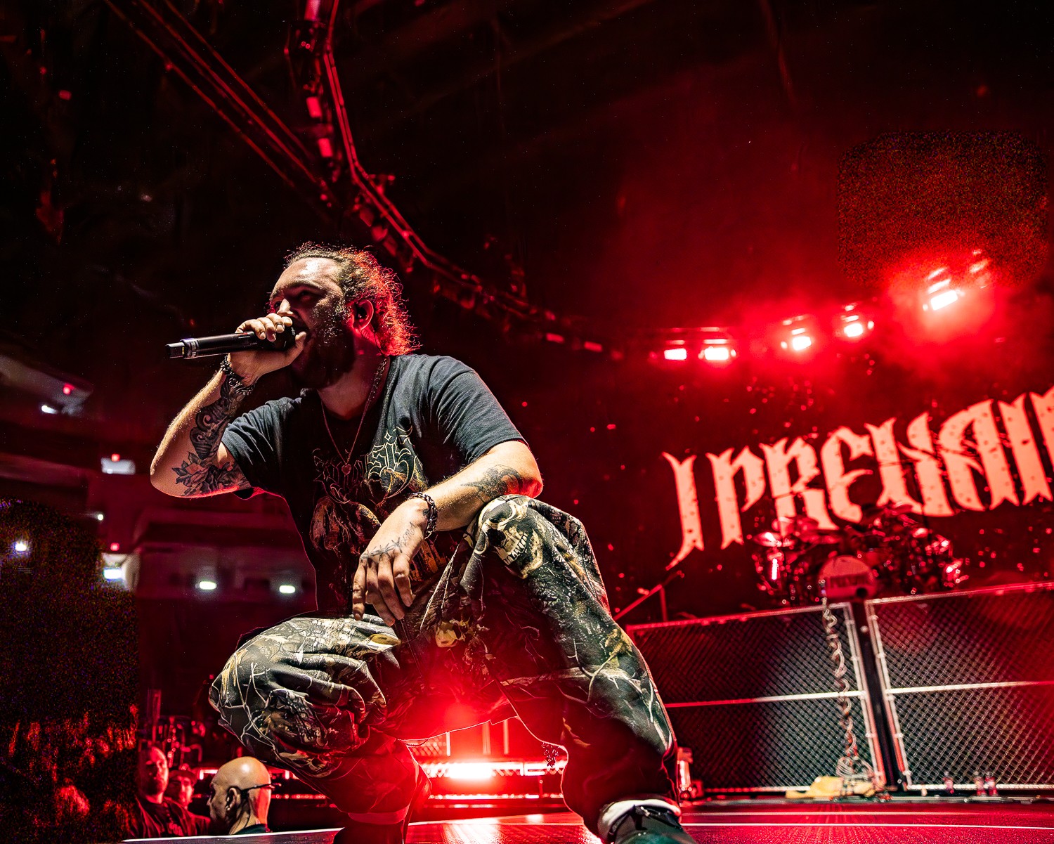 Performer with microphone kneeling on stage, red lighting, and a large screen with text in the background.