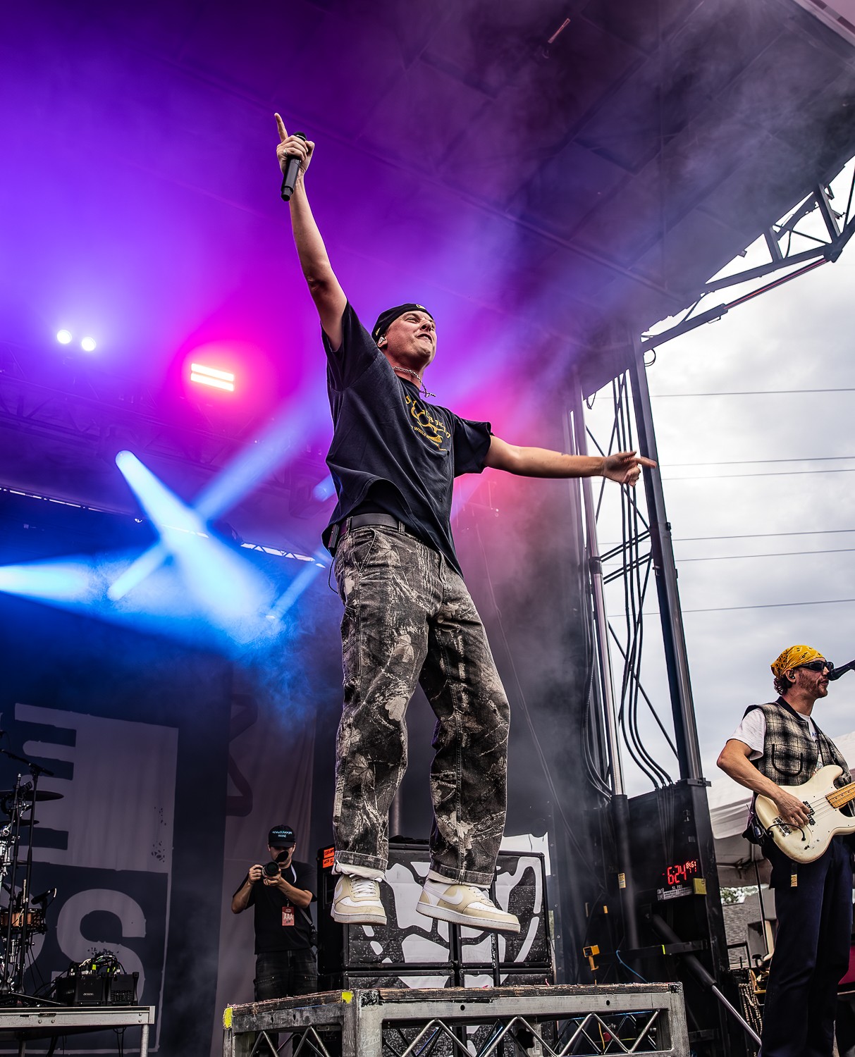 Performer jumps on stage with one arm raised, holding a microphone, with a guitarist and stage lights in background.