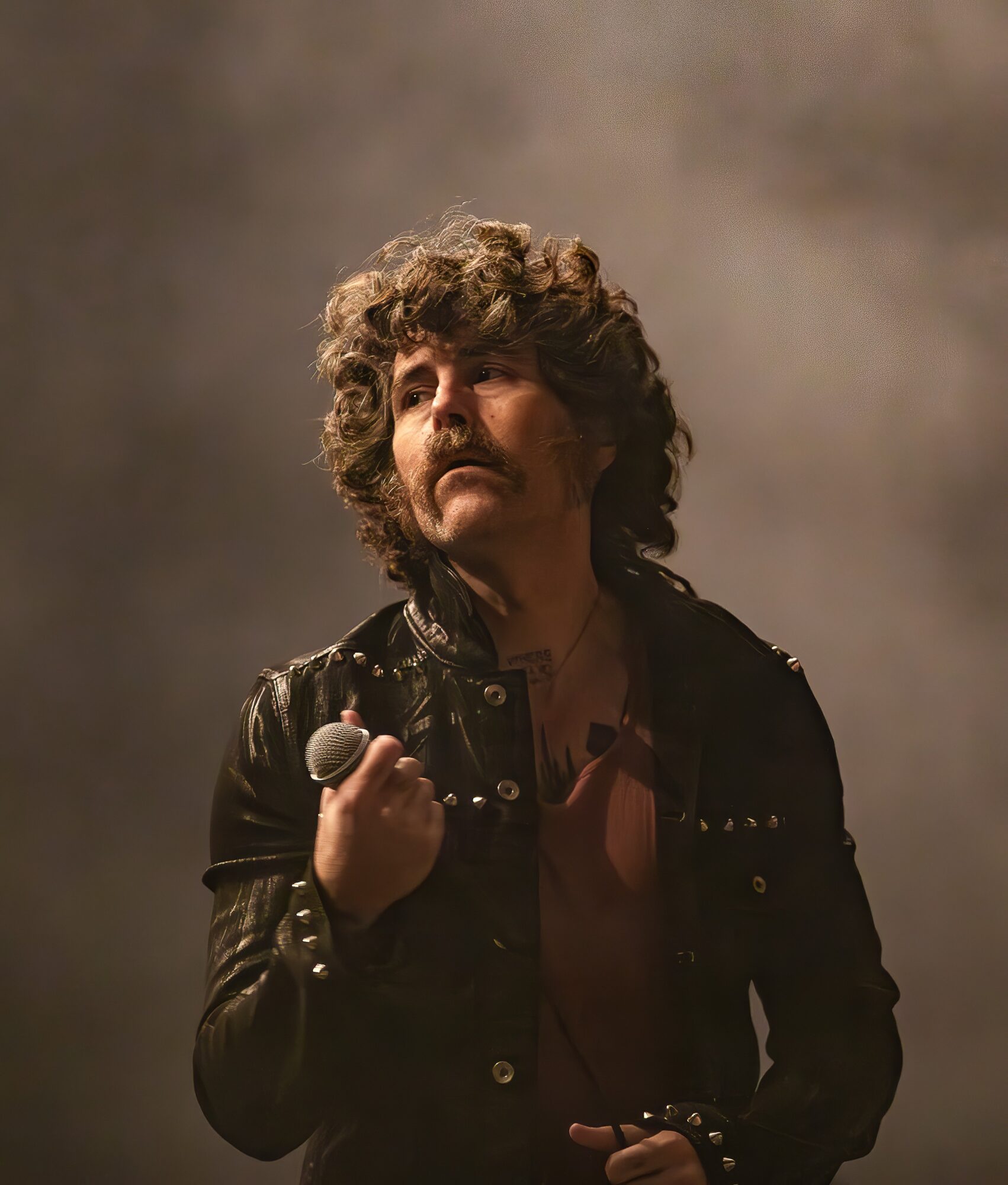 Person with curly hair and mustache holding a microphone, wearing a black leather jacket, on stage with smoky background.