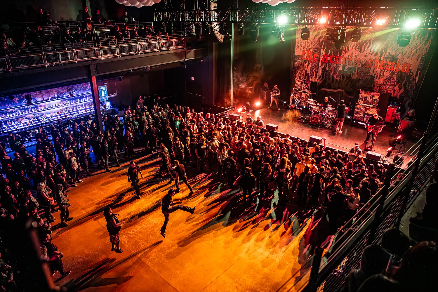 Concert venue with a large audience facing a brightly lit stage, colorful lights, and a mural backdrop.